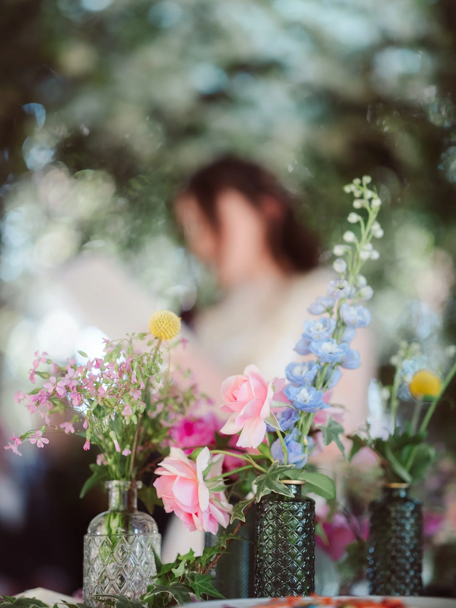 Piccoli vasi in vetro con fiori rosa, azzurri e gialli in primo piano a fuoco con la figura della sposa sfocata sul fondo