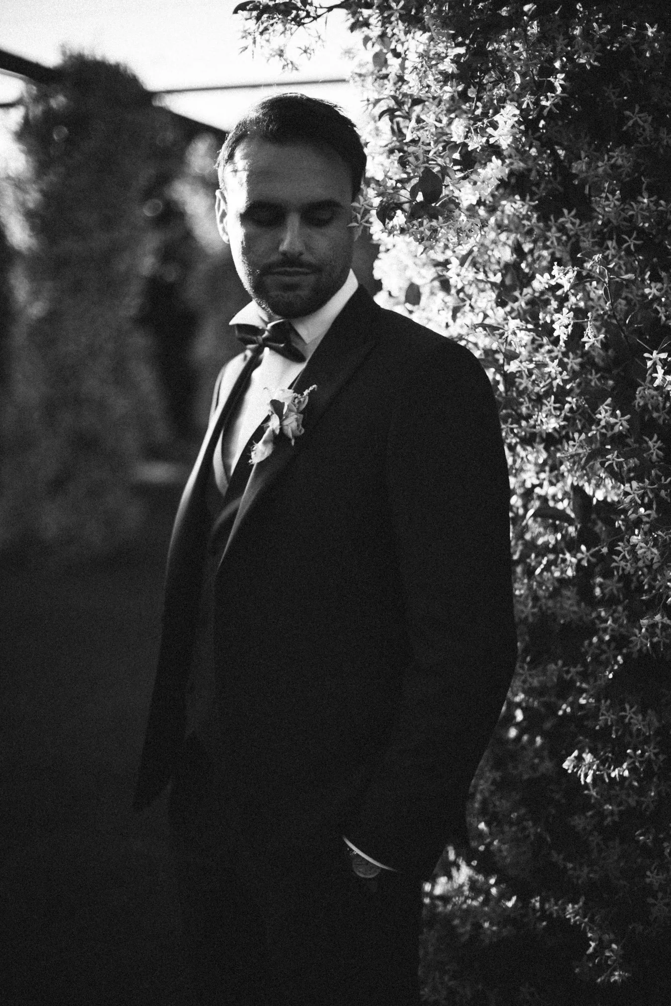 Ritratto in bianco e nero dello sposo in tuxedo elegante con papillon, accanto a una siepe fiorita