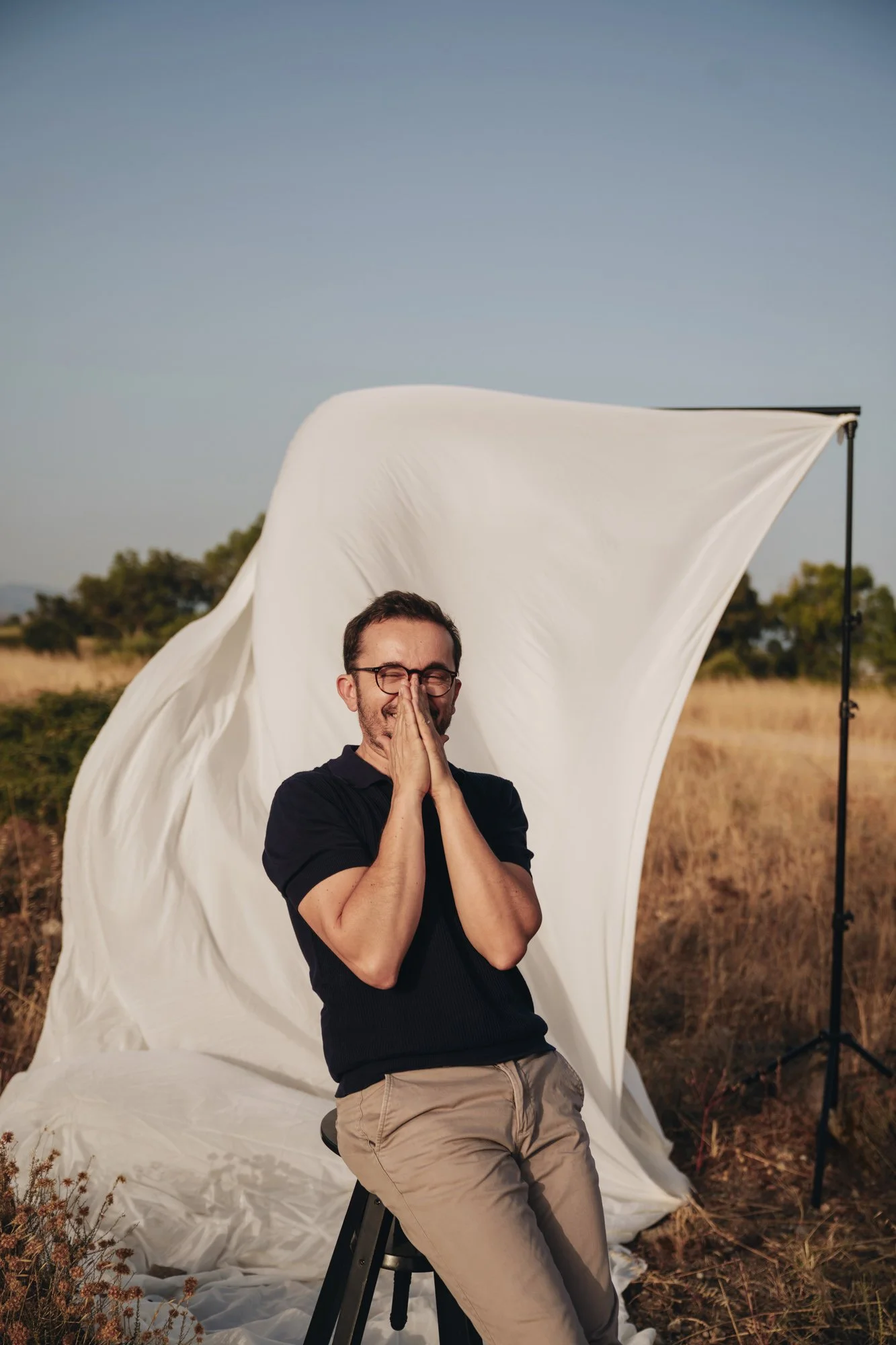 Un uomo sorridente con occhiali si copre il volto con le mani, seduto su uno sgabello in un ambiente all'aperto durante il giorno, con uno sfondo di campagna e un grande tessuto bianco dietro di lui, presumibilmente un set fotografico.