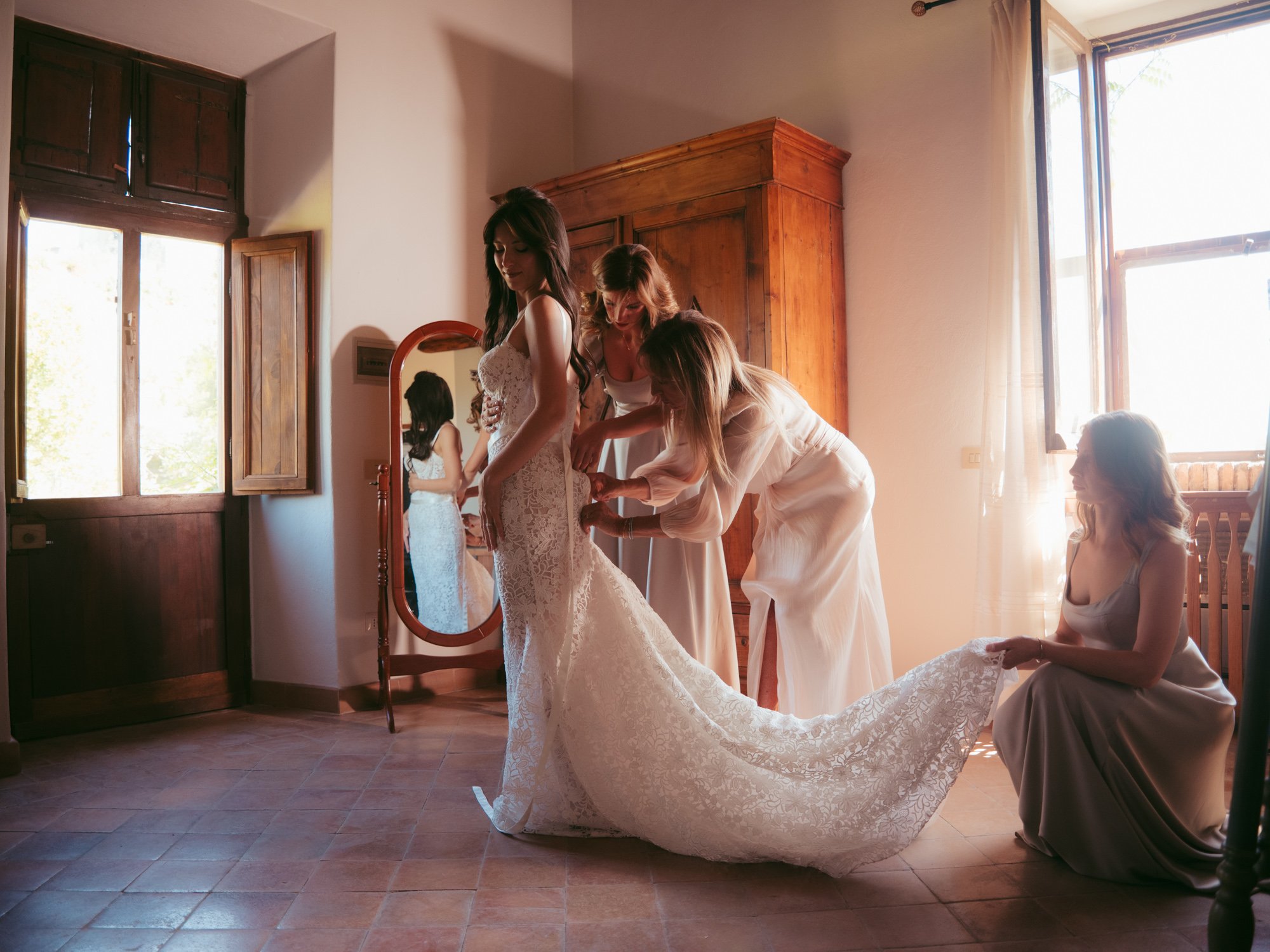 Damigelle e madre aiutano la sposa ad allacciare l'abito in pizzo nella stanza di Borgo di Tragliata