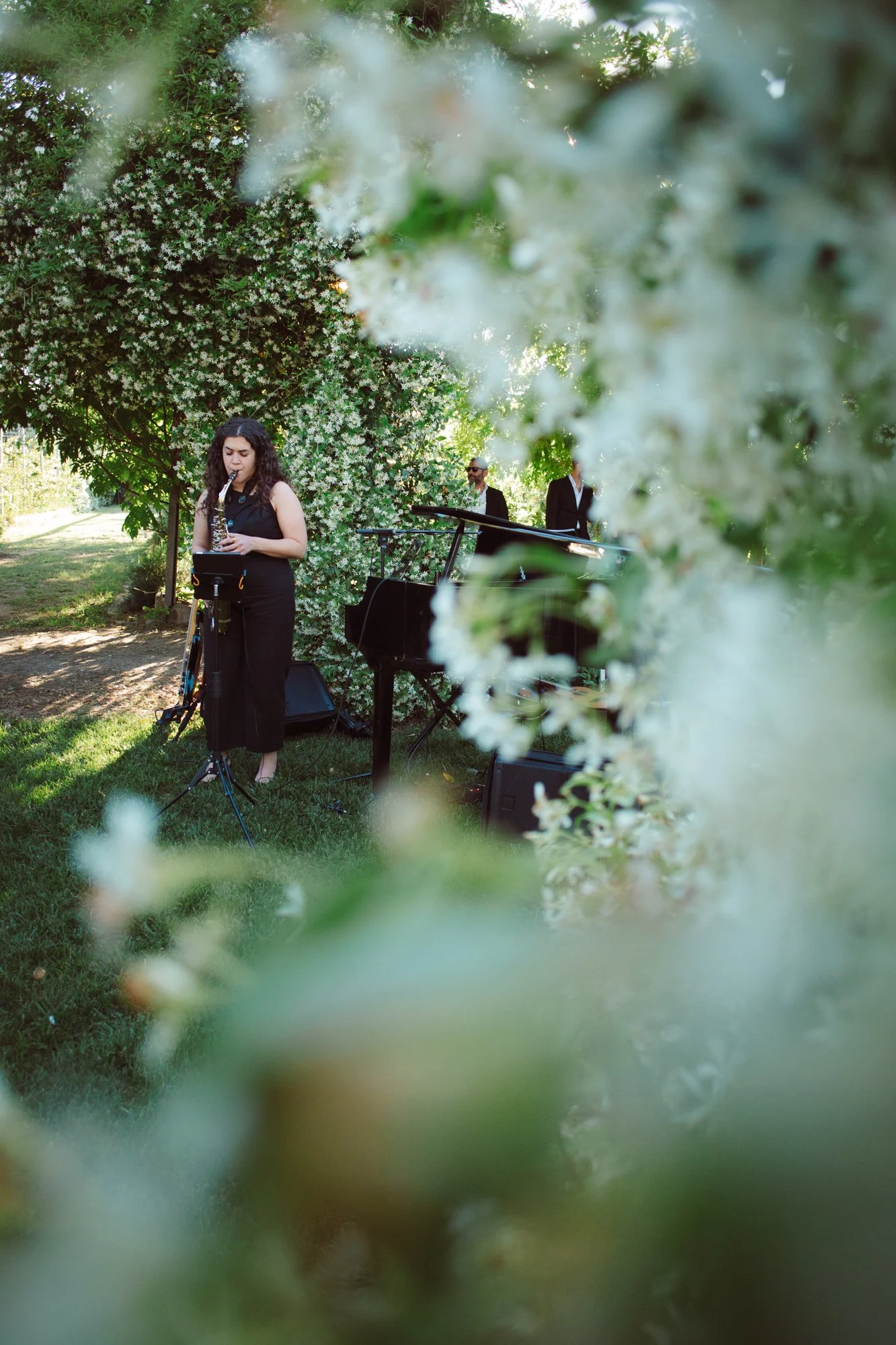 Sassofonista in completo nero che suona il sax nel giardino incorniciata da una siepe di gelsomini in fiore
