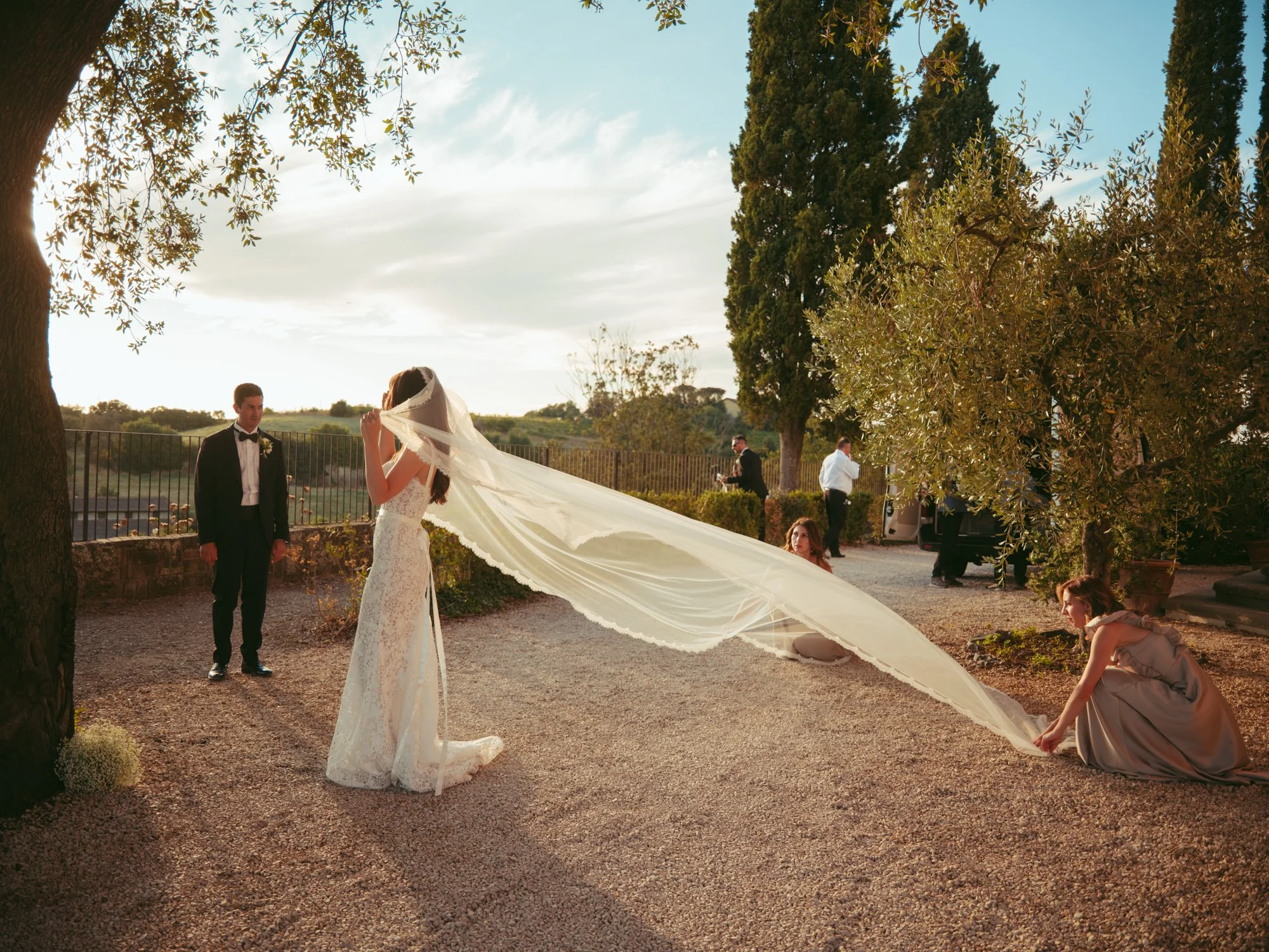 Lo sposo osserva sorridente mentre due damigelle stendono e sistemano il lunghissimo velo della sposa in un cortile alberato al tramonto