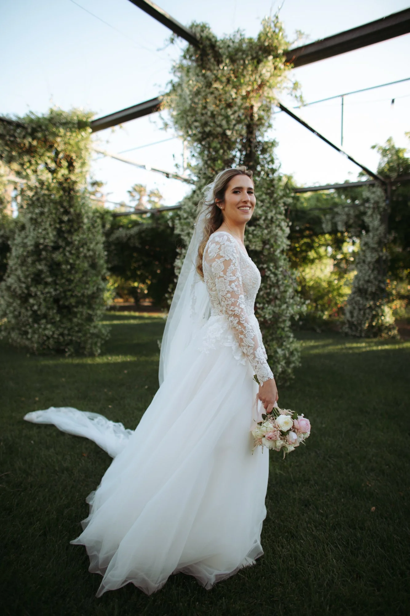 La sposa sorridente in abito bianco con maniche in pizzo si volta nel giardino tenendo il bouquet di fiori rosa e bianchi