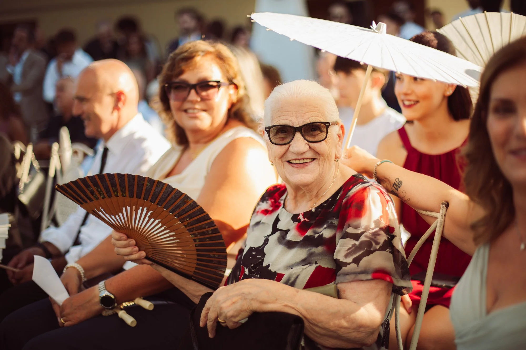Un'affettuosa ospite anziana sorride radiosa tra il pubblico con ventaglio e occhiali da sole alla cerimonia.