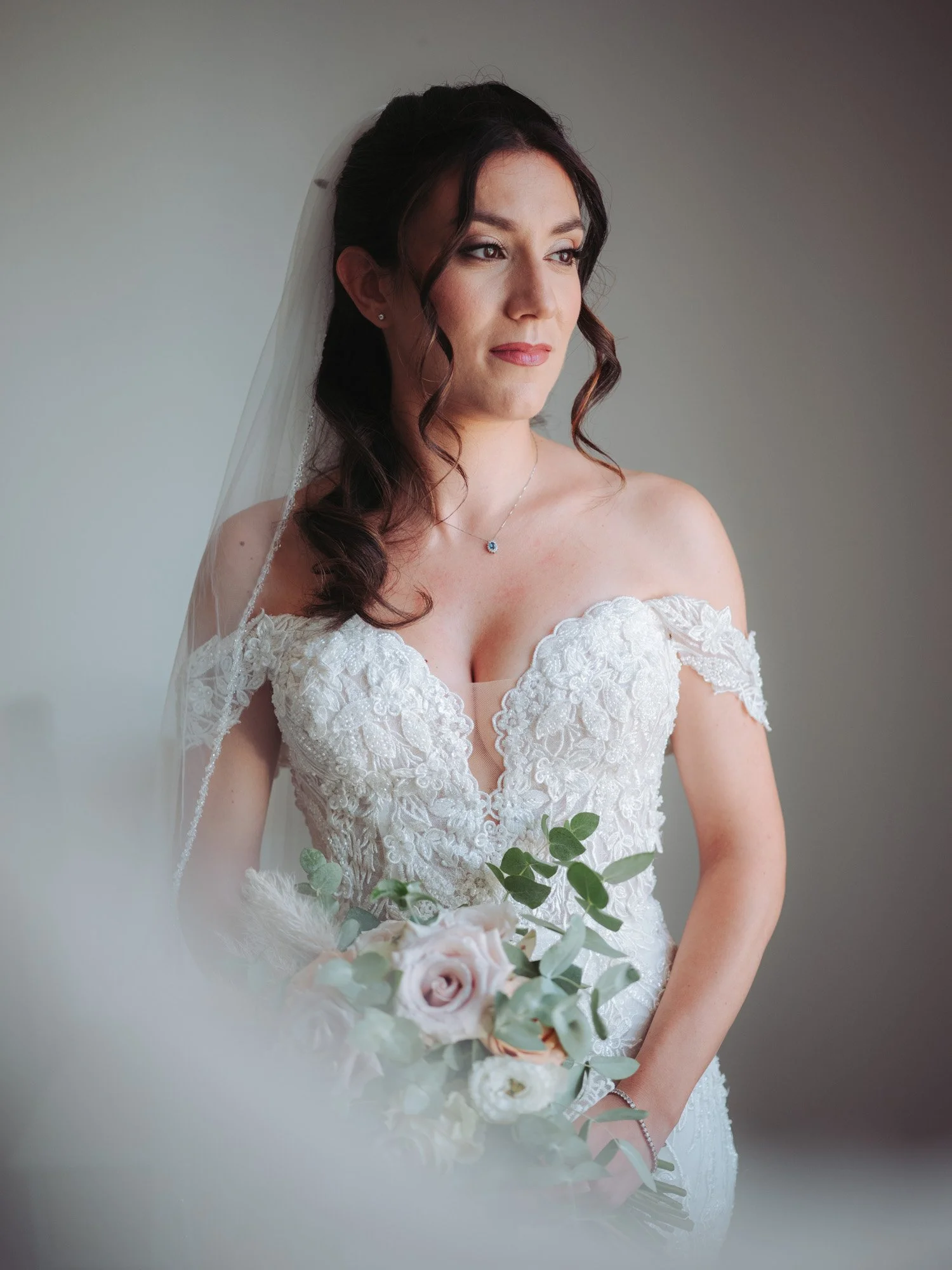 Primo piano della sposa con abito in pizzo, velo ricamato e bouquet di rose nei toni pastello vicino alla finestra