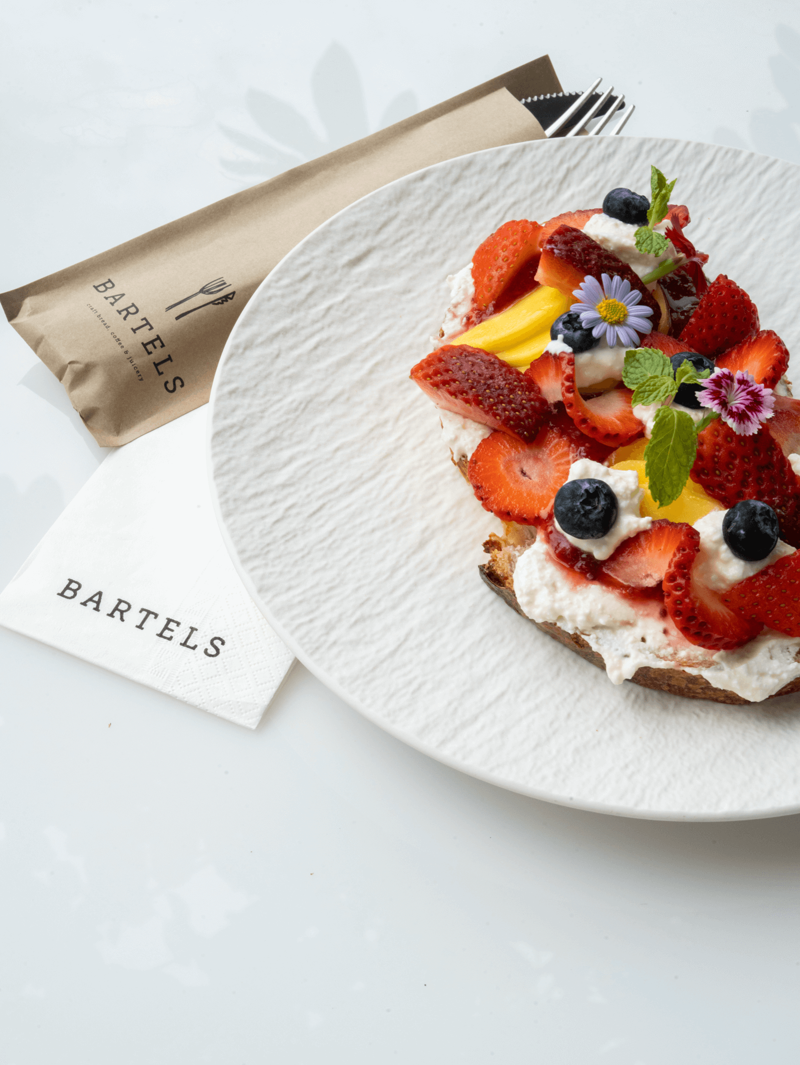 A fruit-topped dessert on a white plate, with strawberries, blueberries, yellow fruit, whipped cream, edible flowers, and mint leaves, beside a folded napkin with the logo 'Bartels' and a fork wrapped in a paper sleeve.