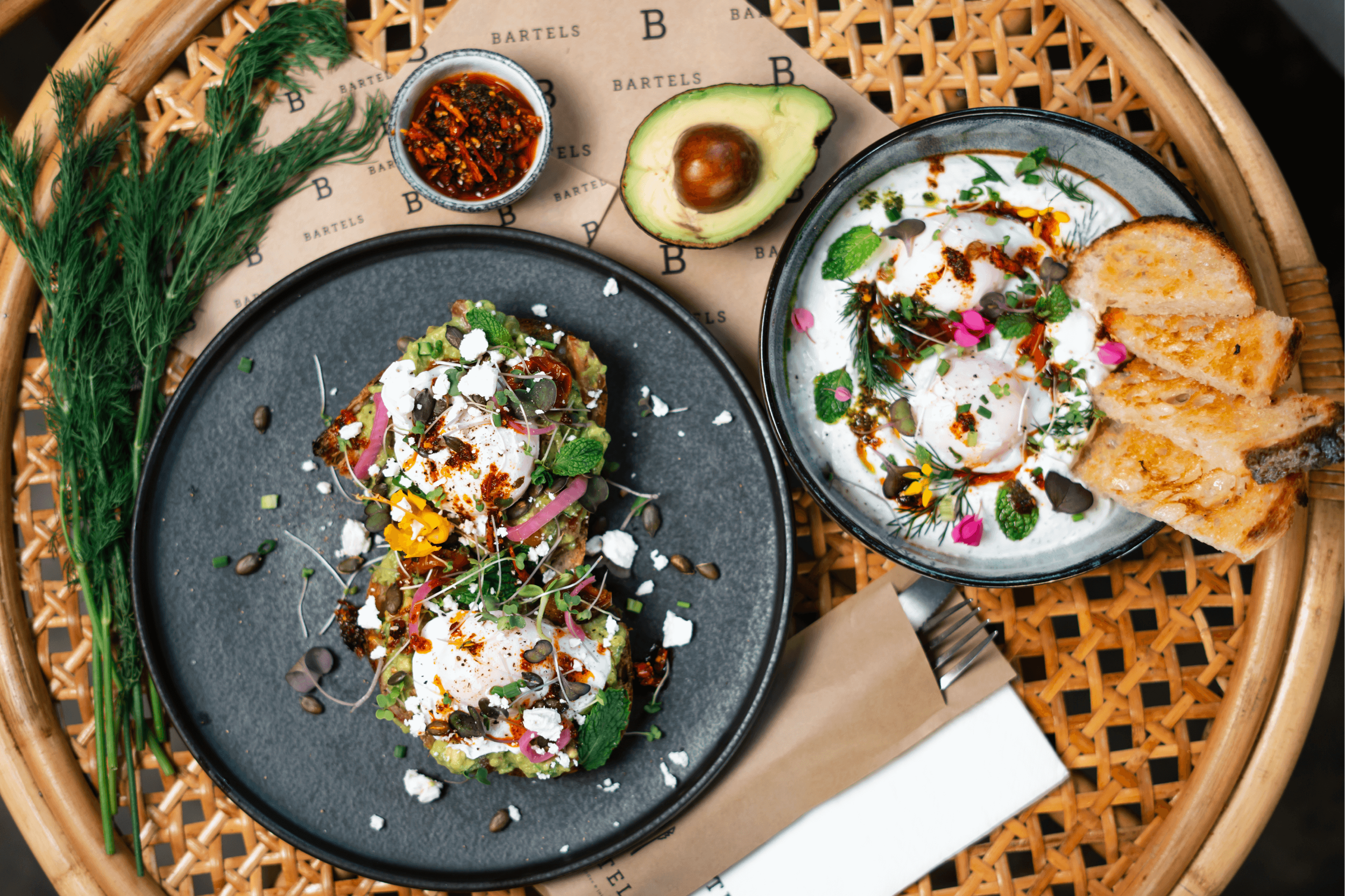 A top-down view of a woven rattan tray holding two dishes of food, a small bowl of sauce, and a halved avocado, with sprigs of green herbs on the side.
