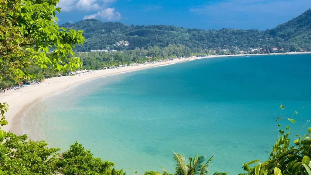 A scenic view of a crescent-shaped beach with white sand, turquoise water, green trees, and mountains in the background.