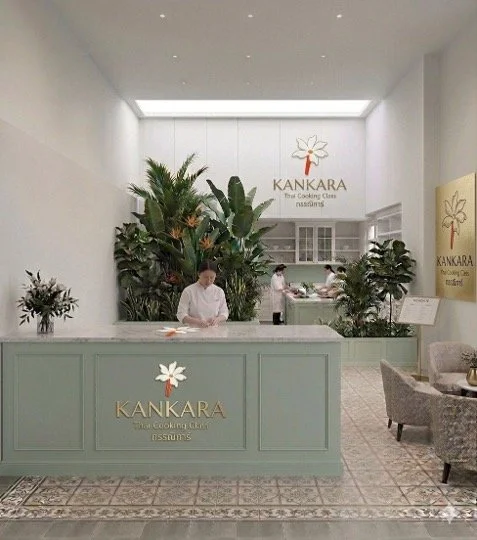 Interior view of Kankara Thai cooking class restaurant, featuring a reception desk with a staff member, lush green plants, and a waiting area with chairs and a table.