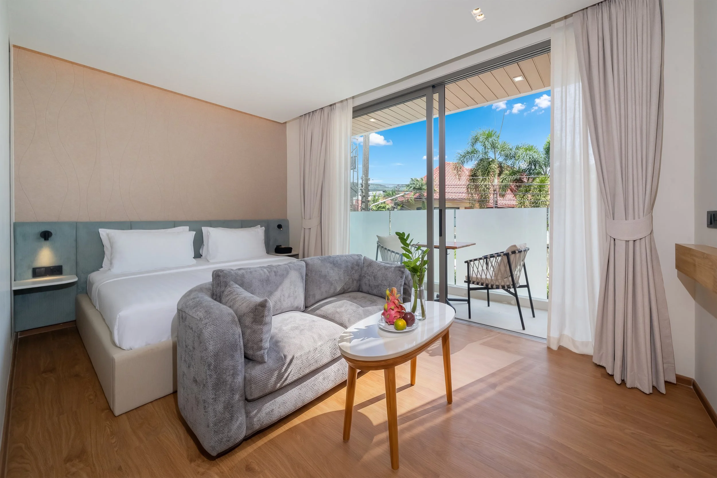 A modern hotel room with a large bed, a gray upholstered sofa, a small round white coffee table with a fruit bowl, a balcony with two chairs, and a view of palm trees and houses under a blue sky.