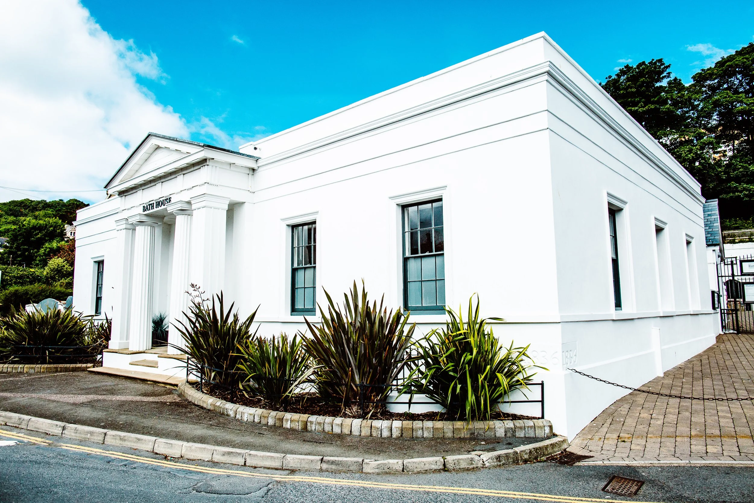 White bathhouse with classical columns and a sign reading 'BATH HOUSE' above the entrance, surrounded by landscaped plants and located on a street with houses and trees in the background.