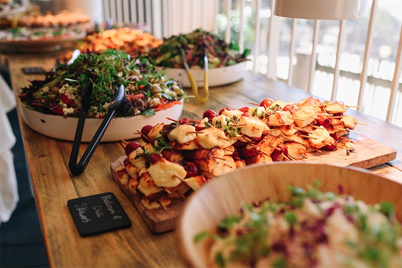 A wooden table with trays of salads and a platter of skewered appetizers at a buffet.
