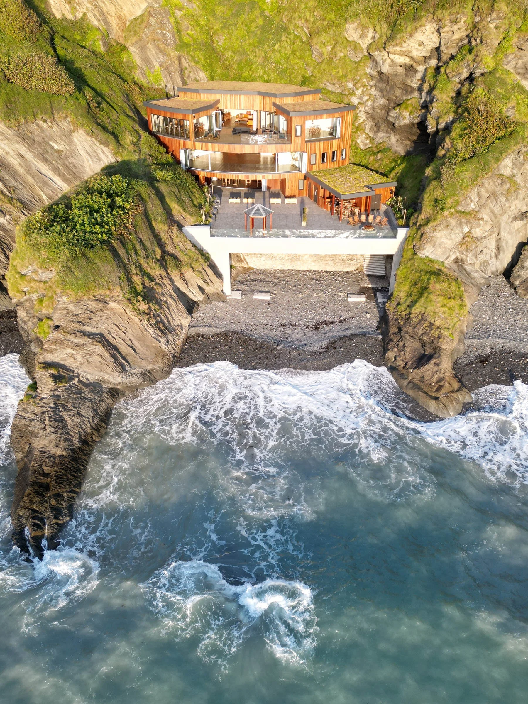 A house built into a rocky cliffside overlooking the ocean with waves crashing below.