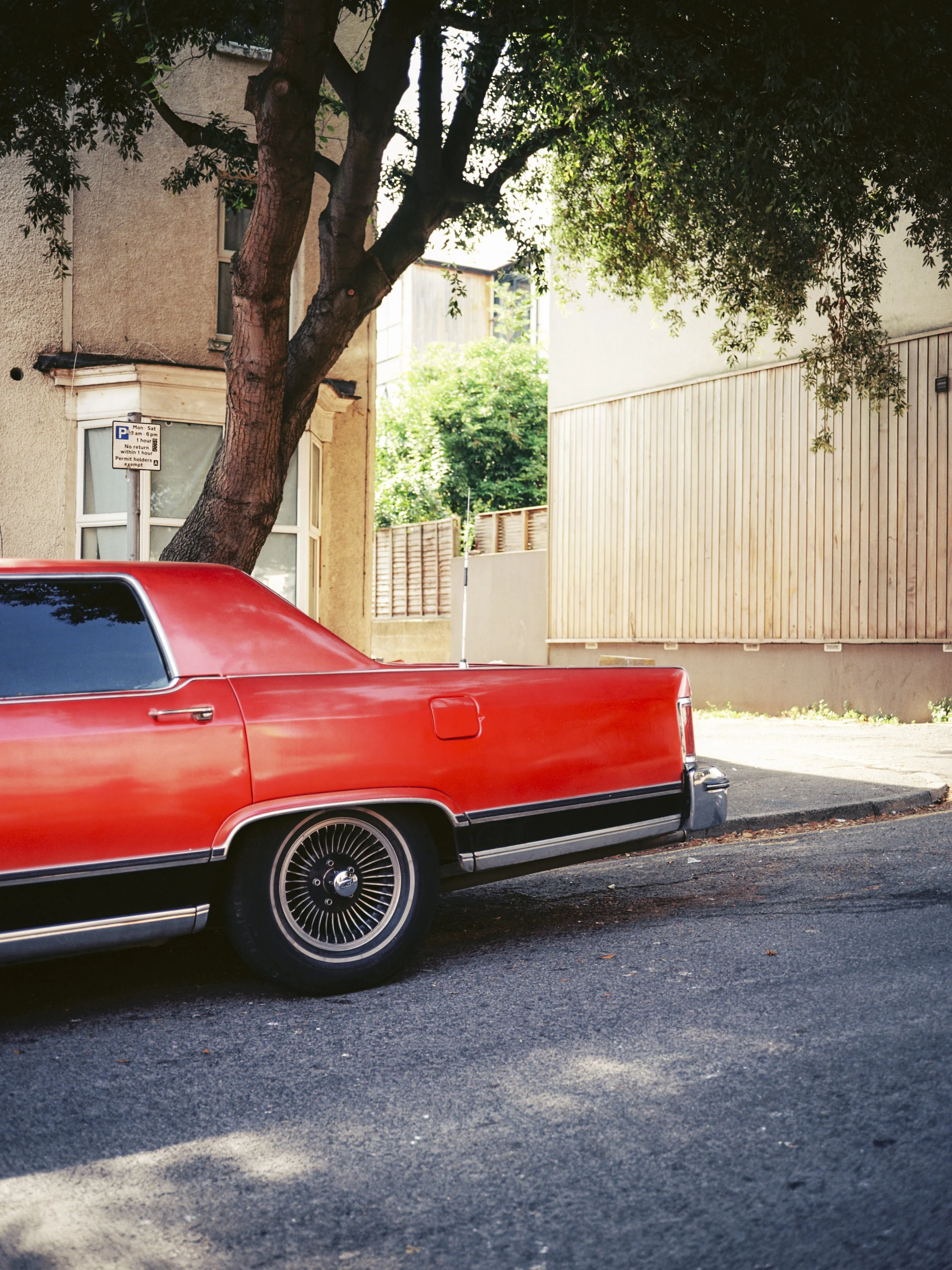 Red vintage car parked along a street with a tree and buildings in the background.