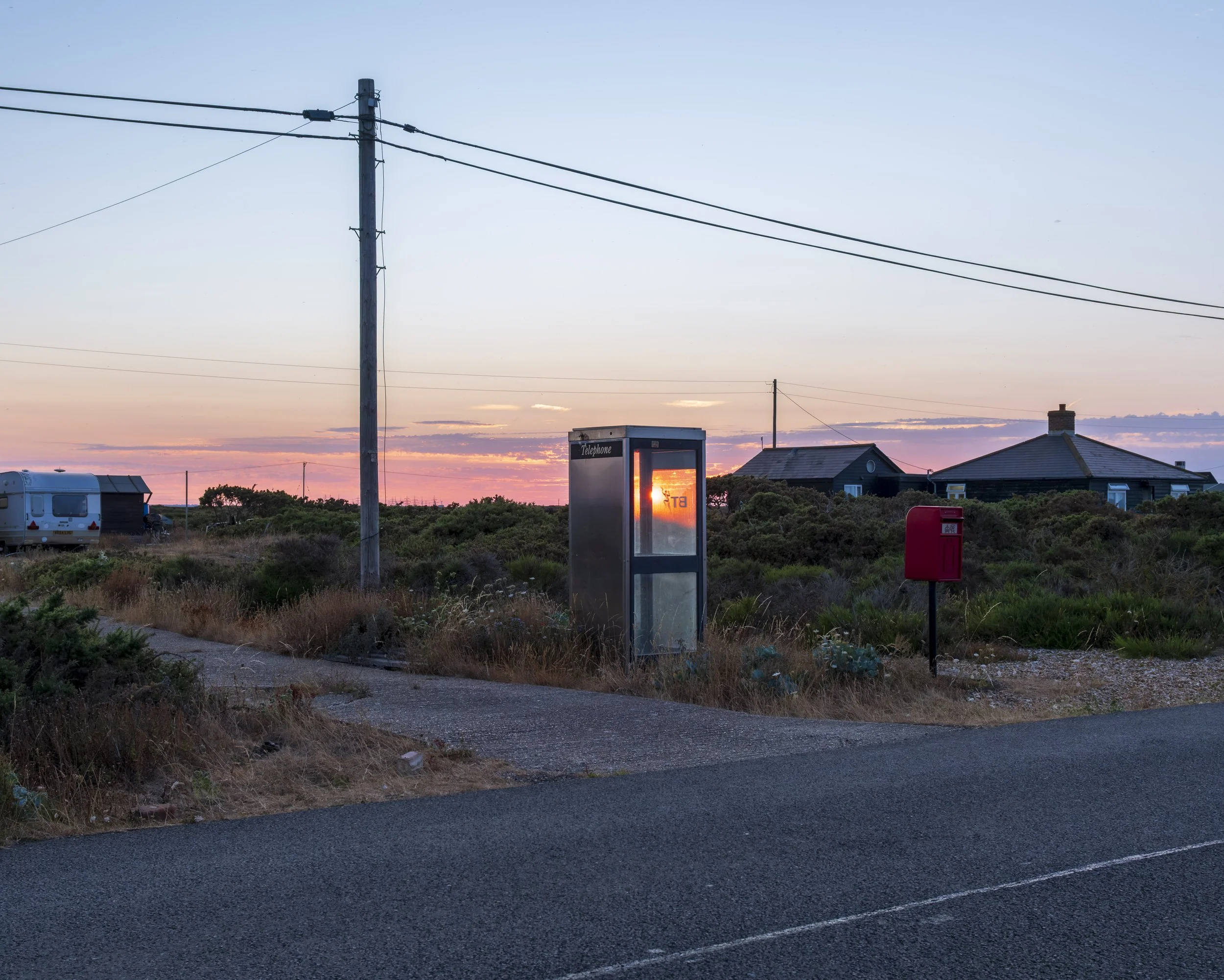 A sunset scene with a telephone booth, a mailbox, a power pole with wires, houses, and a trailer in a rural area with shrubs and a dirt road in the foreground.
