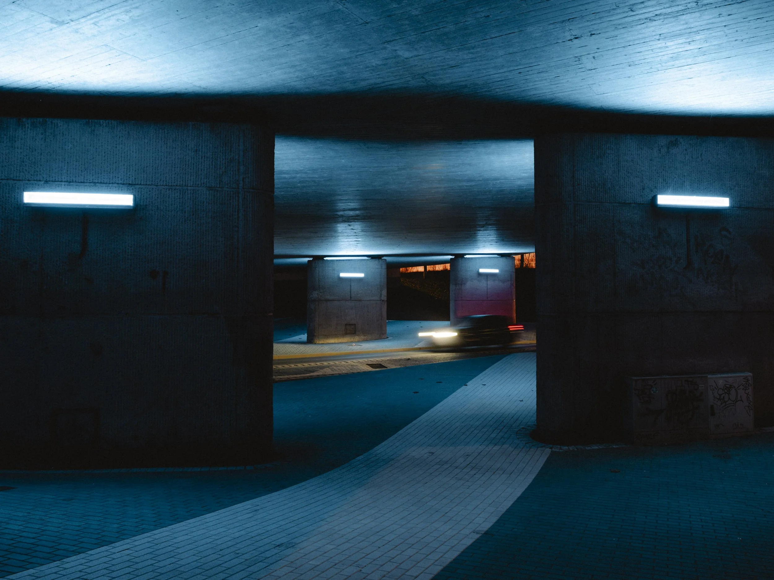 Underpass with blue wall lighting, concrete columns, and tiled floors at dusk.