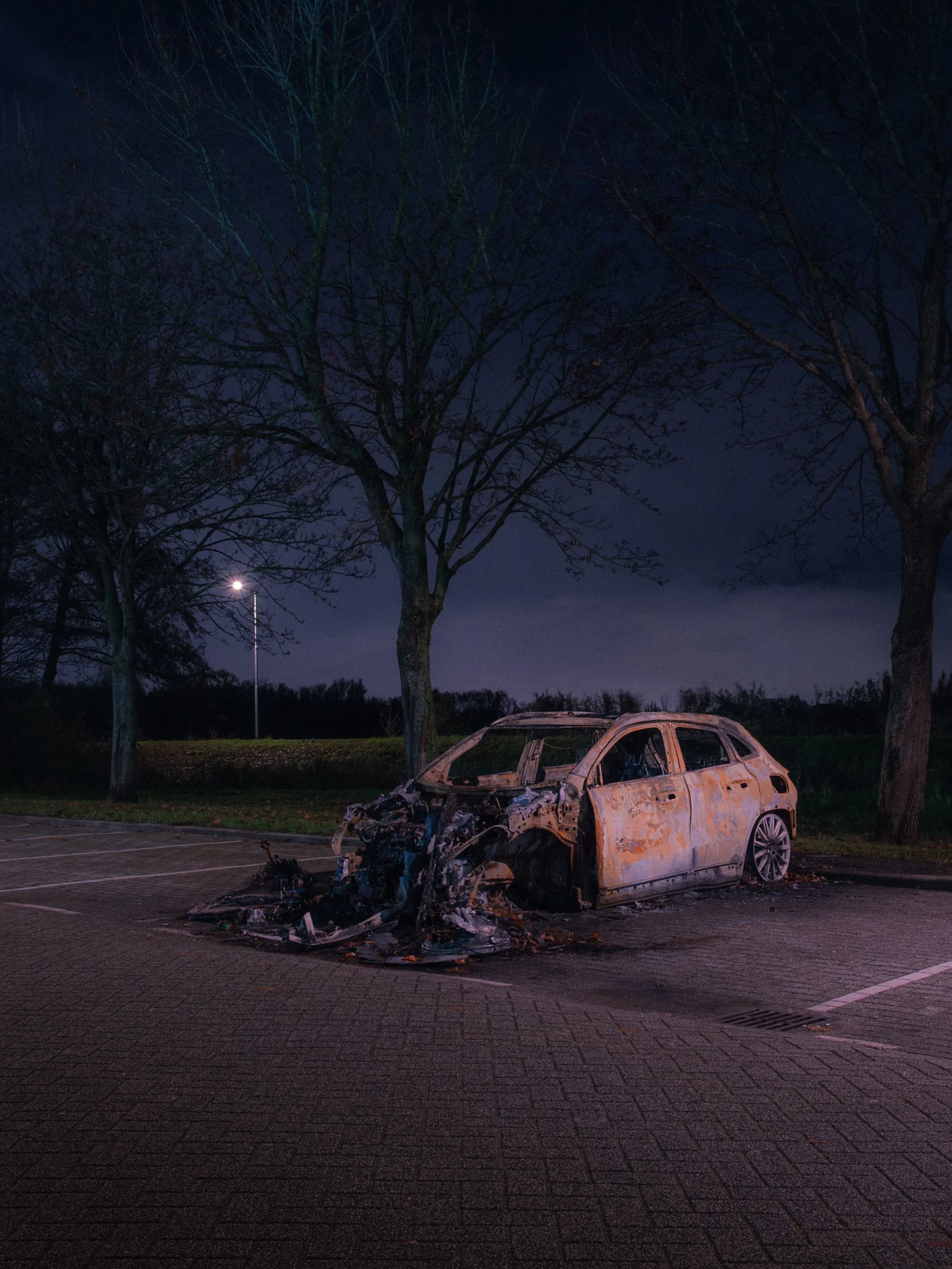 Burned-out and destroyed car in a parking lot at night with trees and a streetlamp in the background.