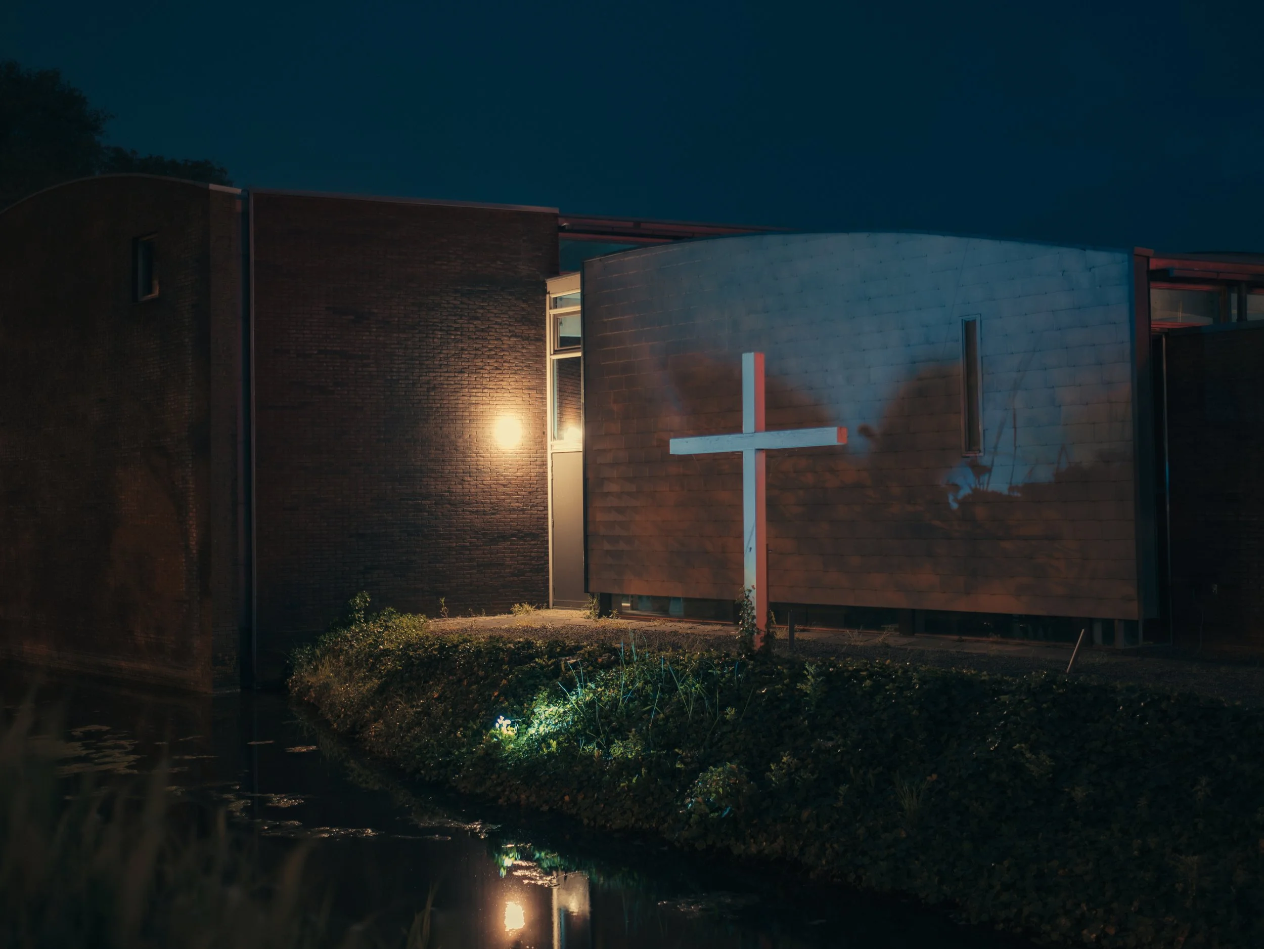 Nighttime scene of a modern church with a white cross, illuminated windows, and a small creek reflecting the cross and building lights, surrounded by greenery.