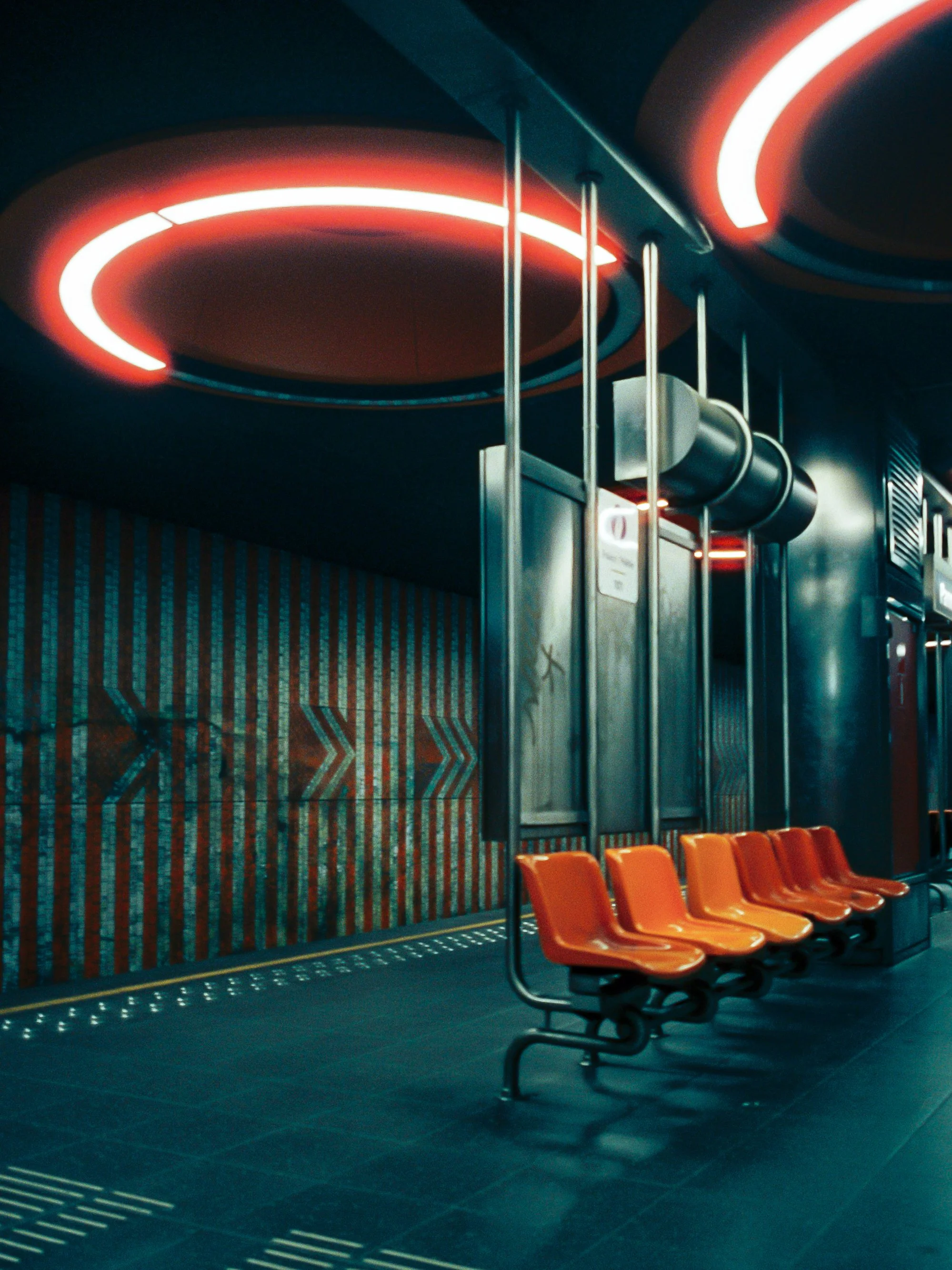Empty orange seats in a waiting area next to metal poles and a wall with colorful lighting on the ceiling in an airport or train station.
