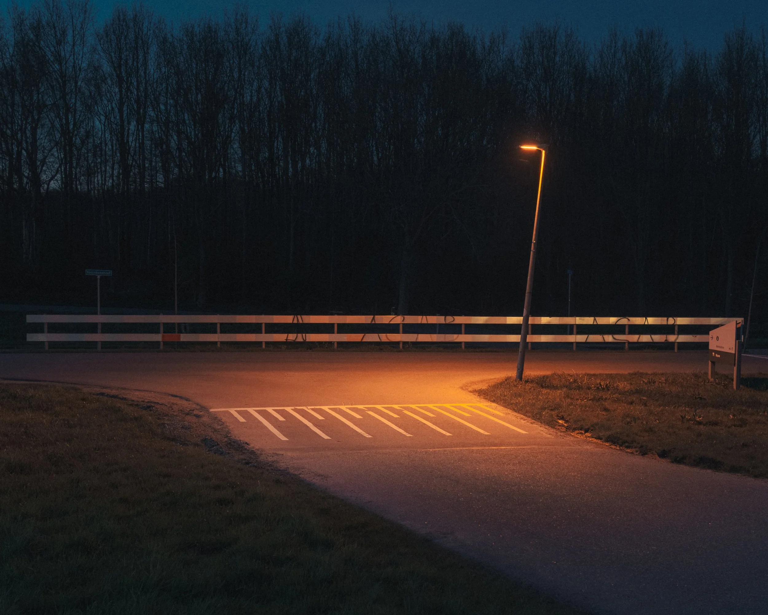 Nighttime scene with a single leaning street lamp casting orange light on the road below, line of leafless trees in the background, and a white fence in the foreground.