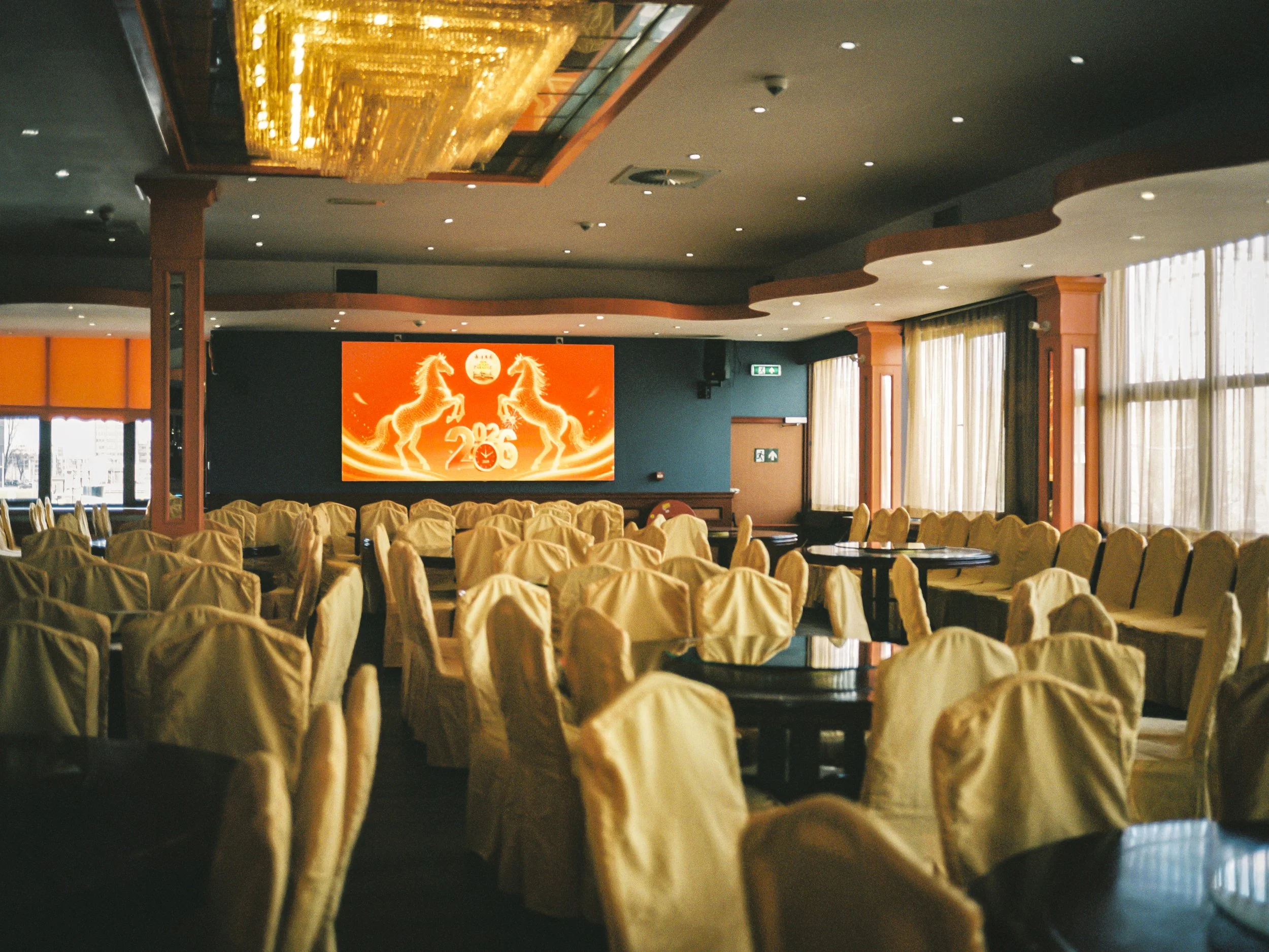 Empty banquet hall with rows of covered chairs, large windows with curtains, and a large digital screen showing gold horses and upcoming event date.