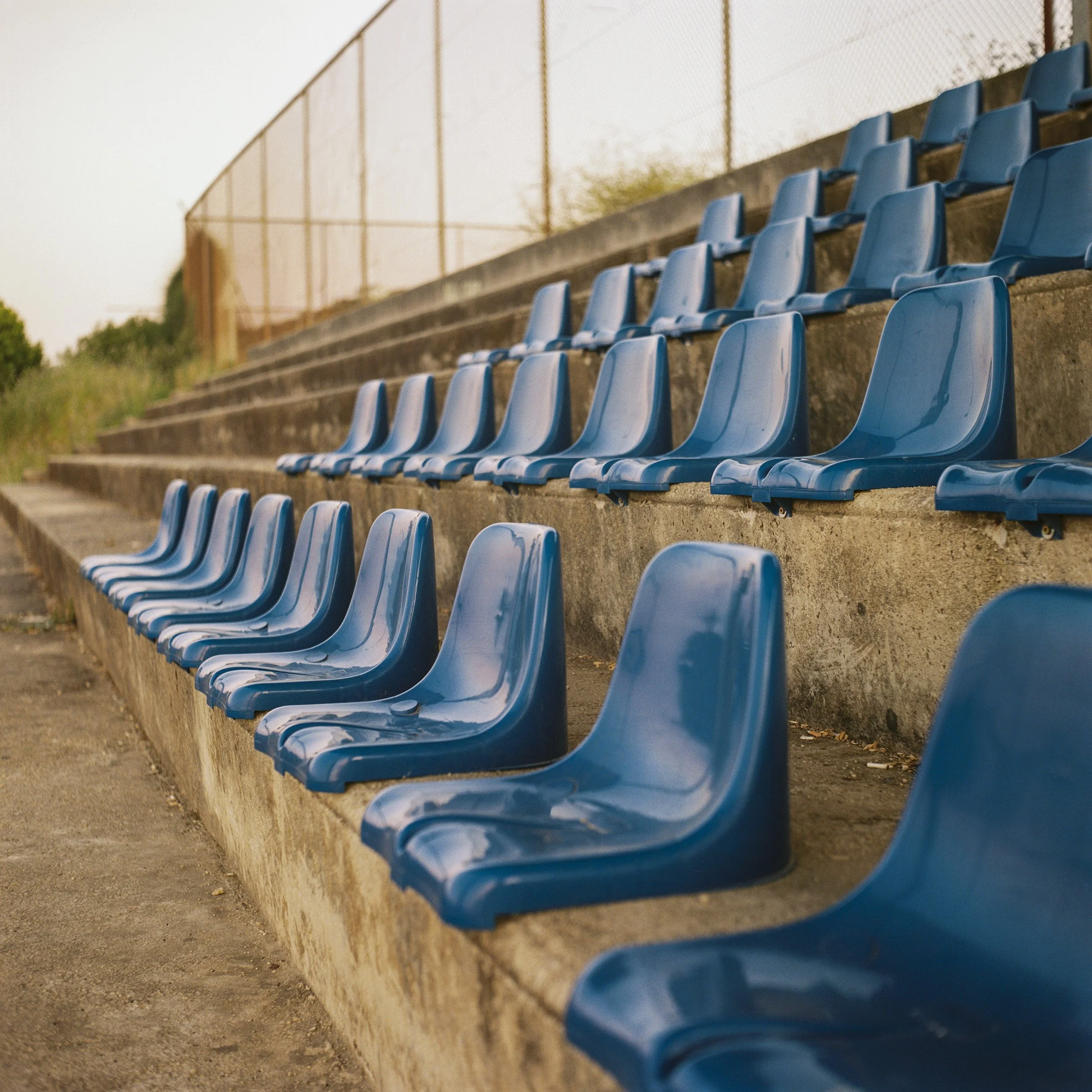 Empty blue stadium seats on concrete steps with a chain-link fence and trees in the background.