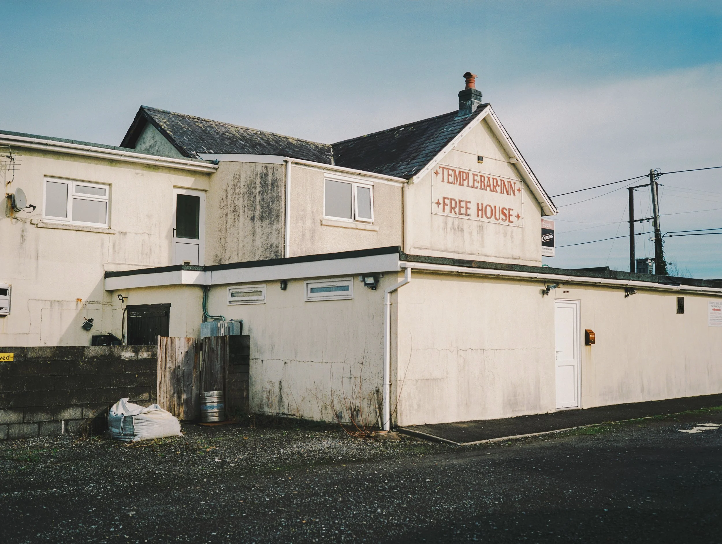 A two-story white building with a sign that reads 'TEMPLATE BAR INN FREE HOUSE' on the upper part of the wall. The building has several windows and a door, with electrical wires and poles visible nearby. The ground in front is gravel with a few items