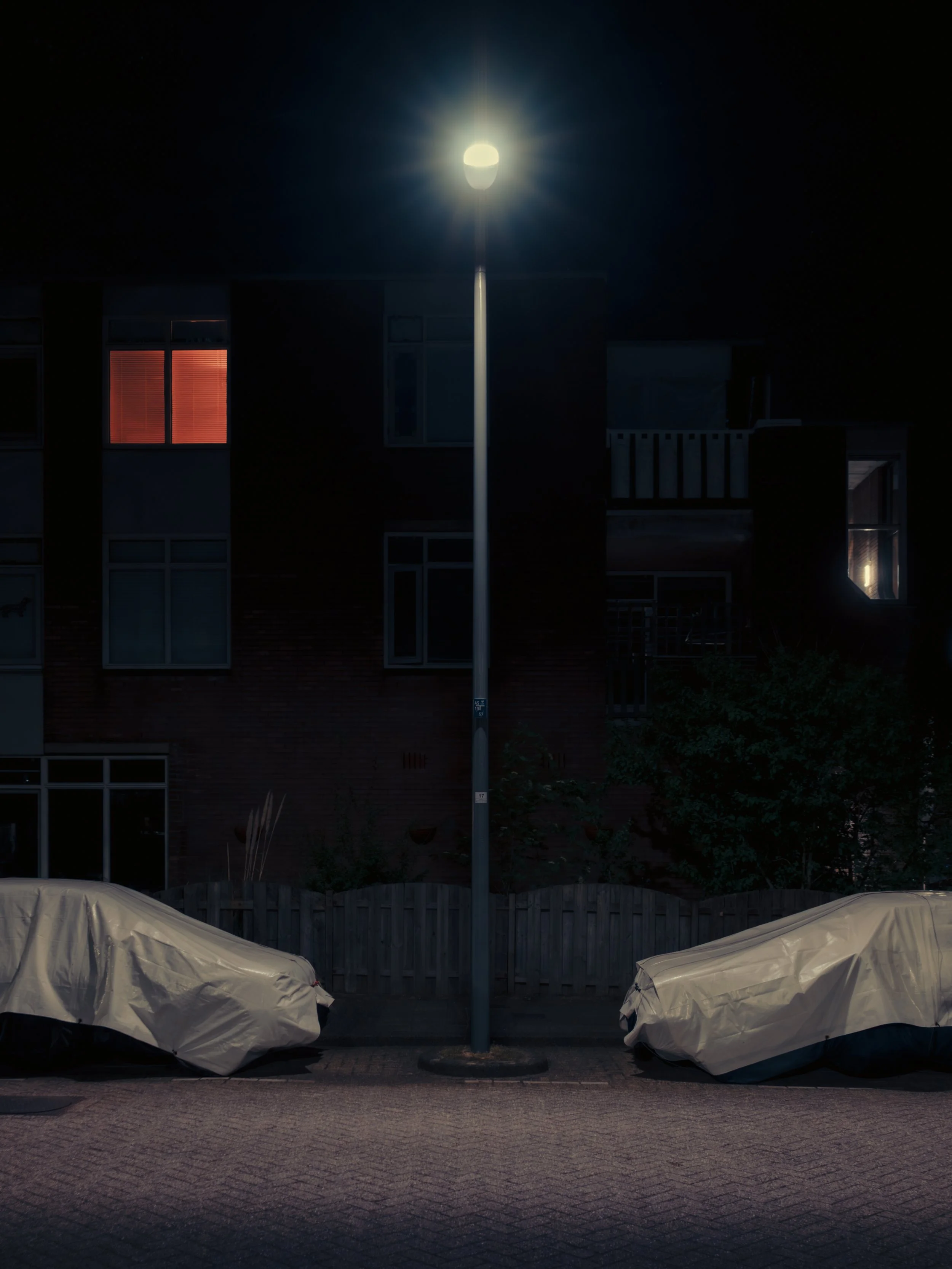 A street at night with a tall streetlamp casting bright light. Two covered objects, possibly boats or furniture, are positioned on the ground in front of a wooden fence. A multi-story building with illuminated windows, including one with glowing red 