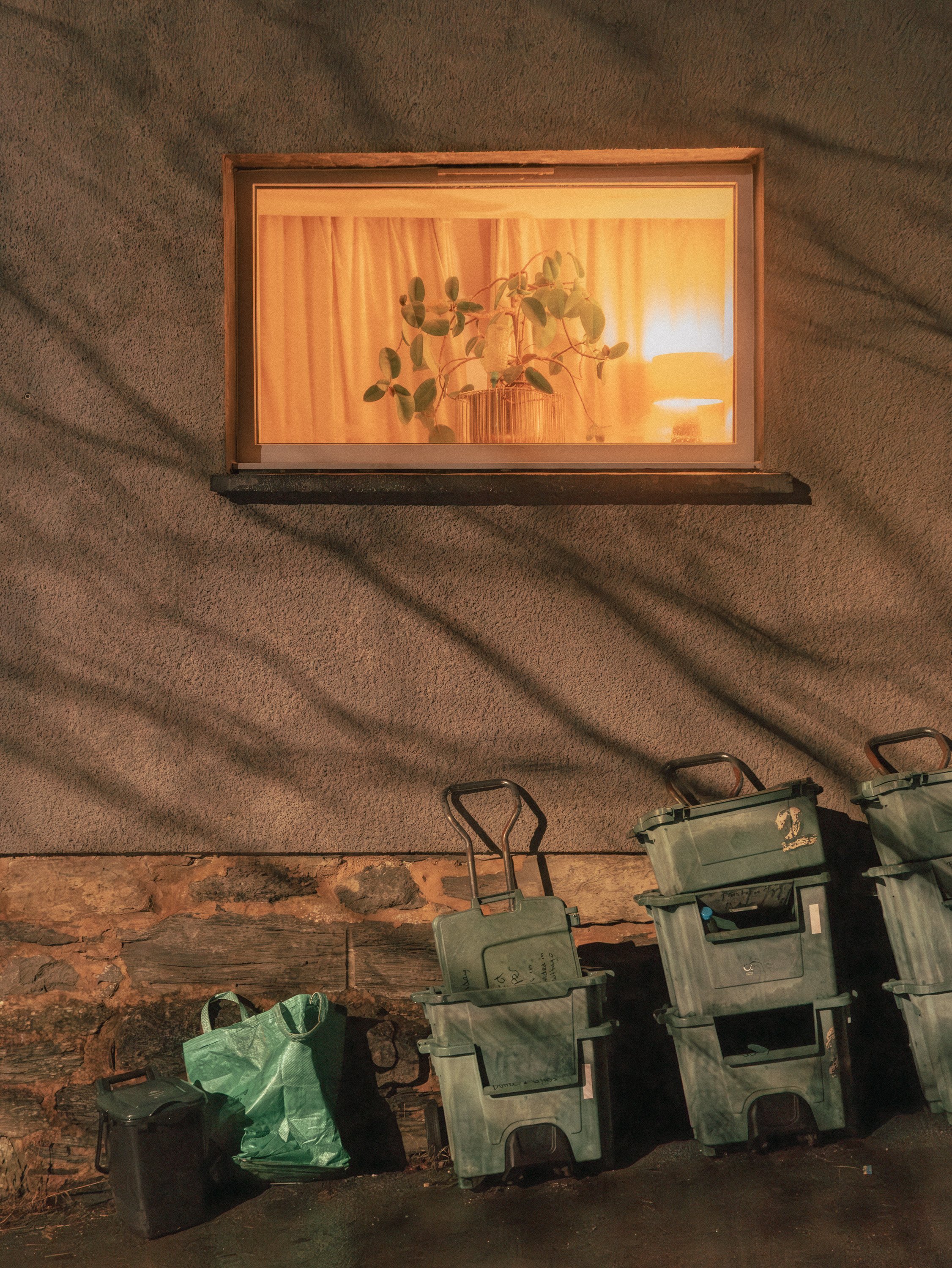 Stacked green and black trash bins on a concrete ground with a brick wall and an illuminated window of a house in the background.