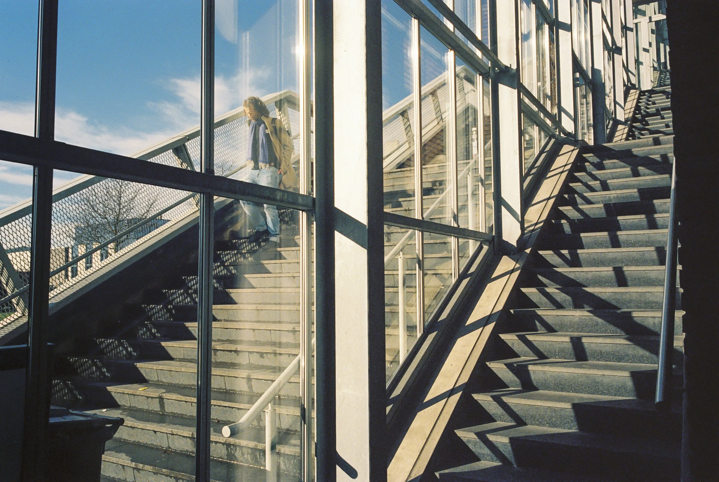 A woman with curly hair wearing a beige coat and backpack walking up an exterior staircase of a modern glass building on a sunny day.