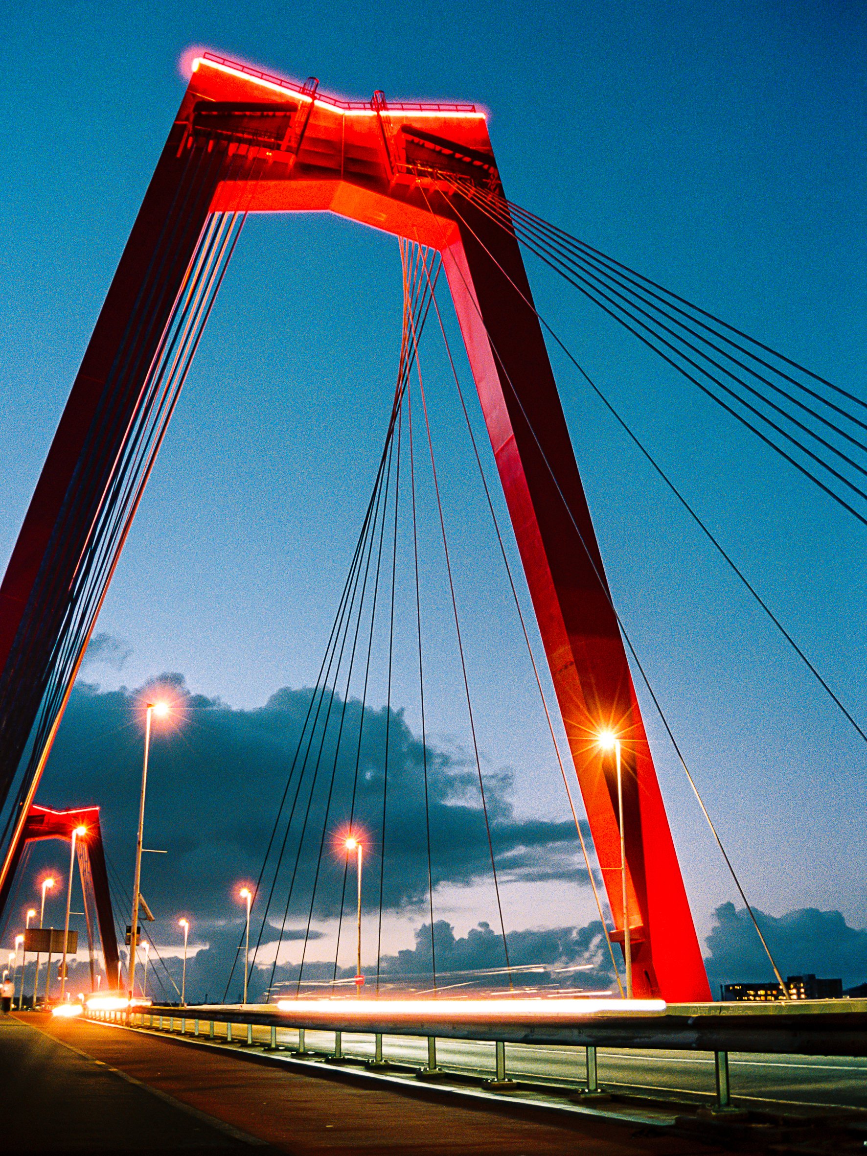 Nighttime view of a modern cable-stayed bridge with a large red arch and illuminated support cables, under a cloudy sky.