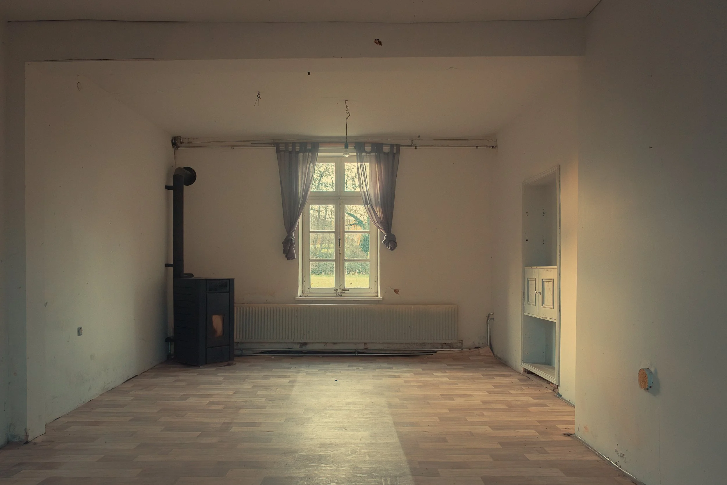 An empty room with a large window, grey curtains, a wood stove, and a radiator underneath the window. The walls are white and slightly worn, with some visible marks and holes. The flooring is light-colored wood. There is an open wall space on the rig