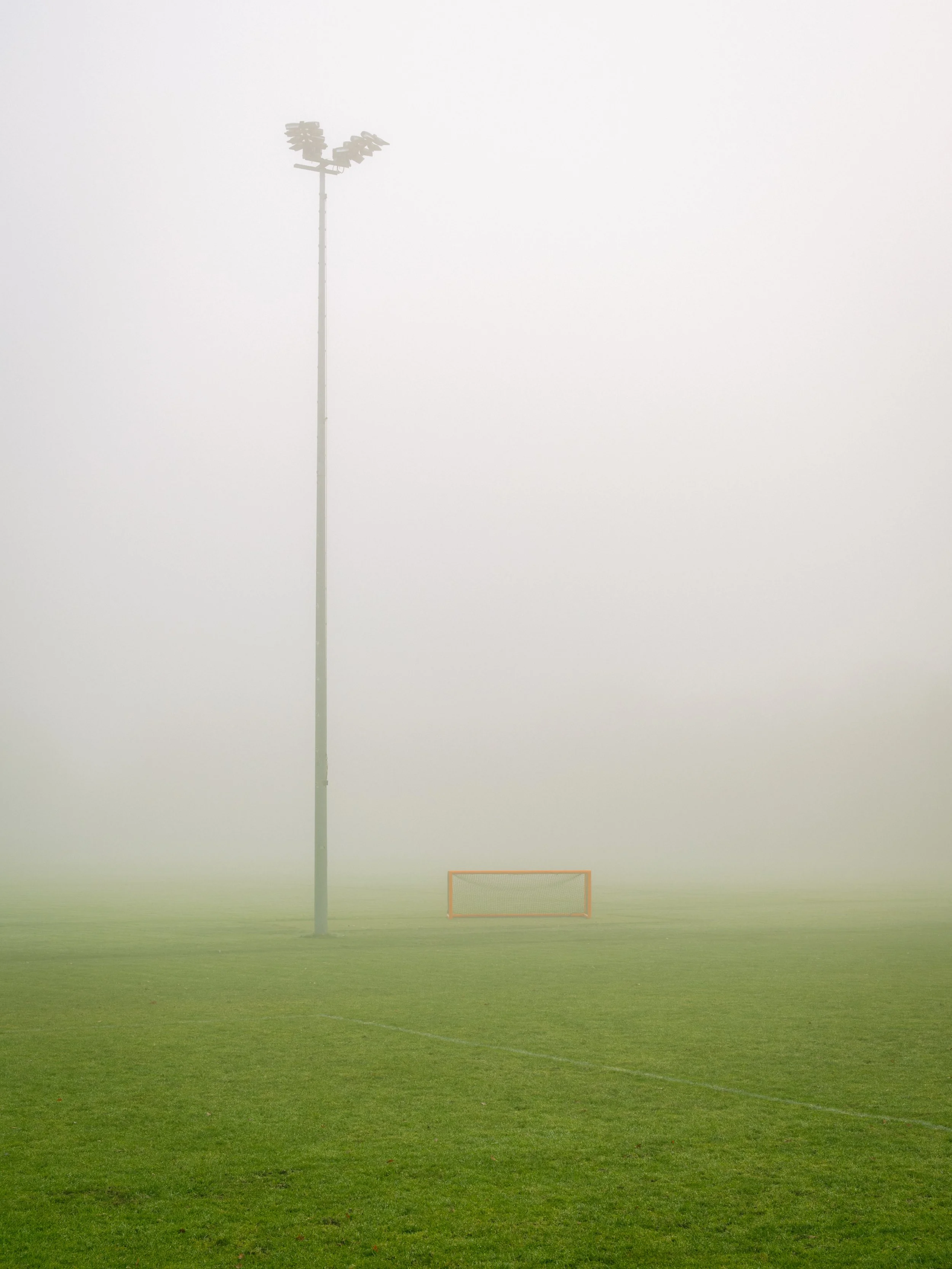 An outdoor sports field with a tall floodlight pole and a small goal post, all shrouded in fog.