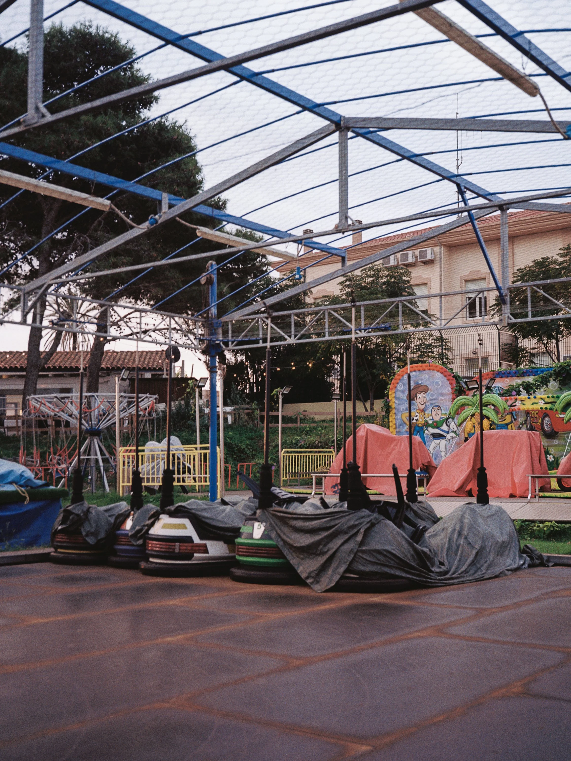 The image shows an amusement park or carnival ride area with a partially assembled or dismantled ride structure made of metal at dusk. There are happiness-themed children's rides with cartoon characters, colorful vehicles, and stalls covered with tar