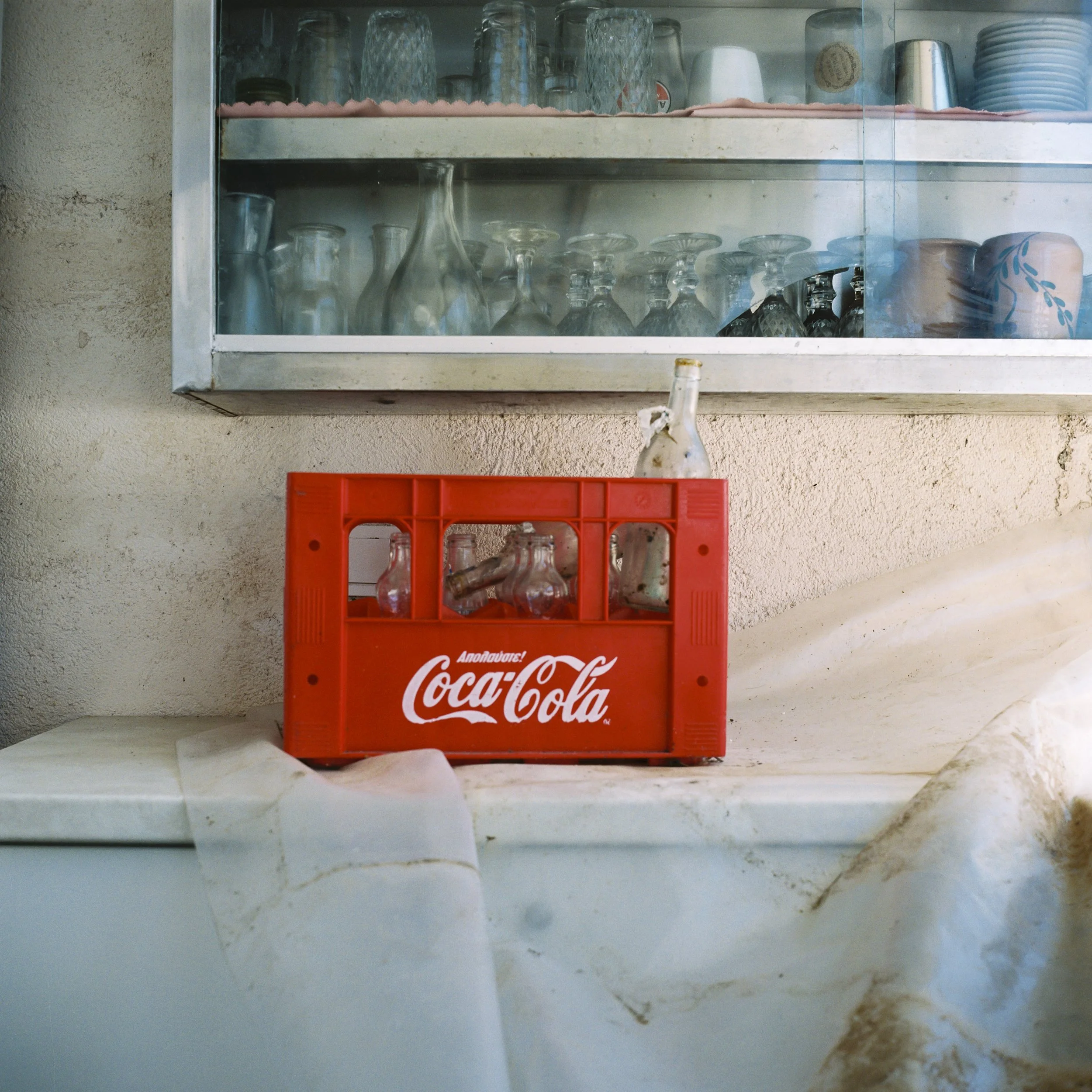 A glass display case mounted on a wall containing various clear glass cups and bottles, with a red Coca-Cola branded crate with bottles placed below.