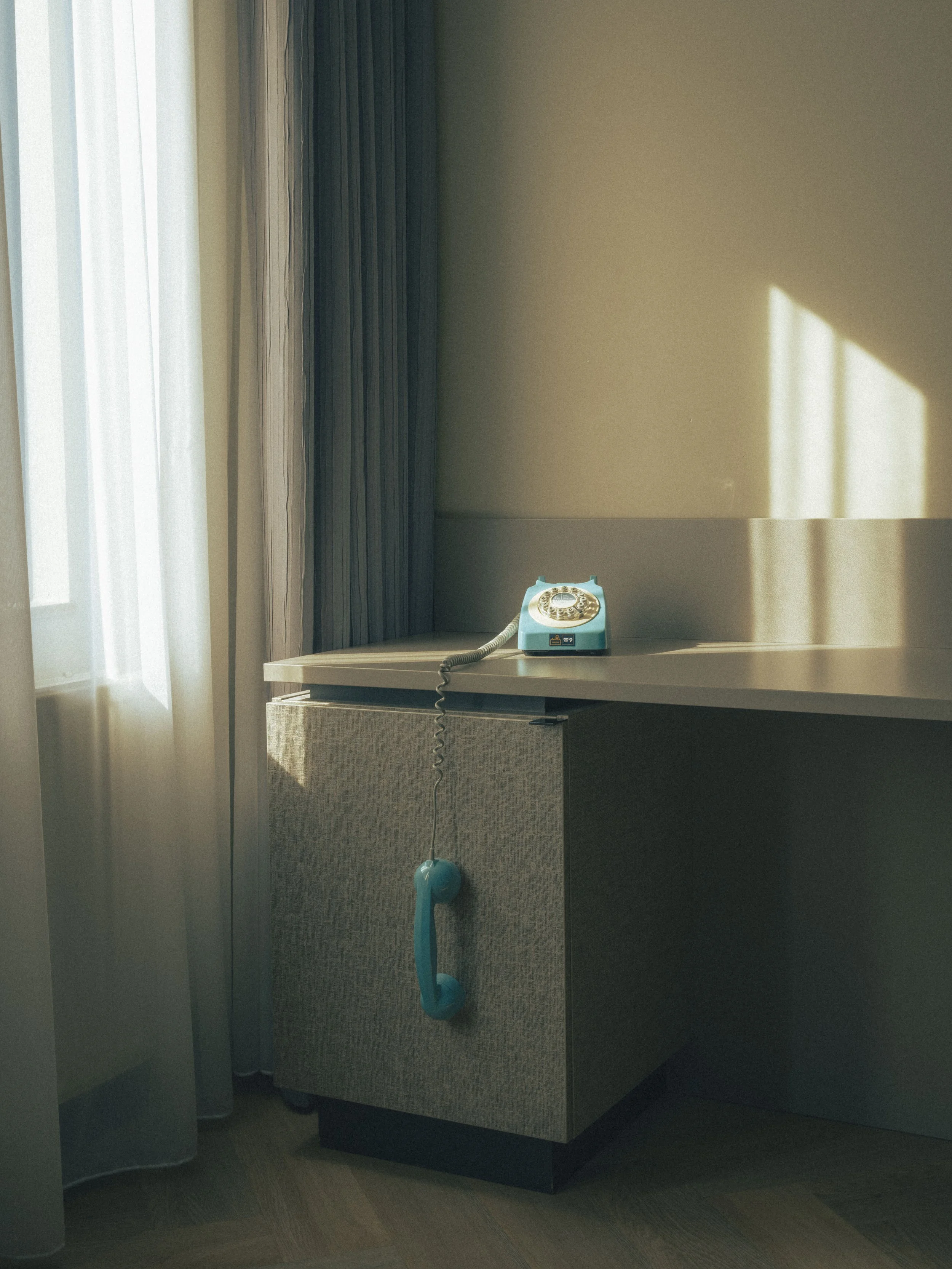 A hotel room corner with a beige wall. An old-fashioned blue rotary phone sits on a beige cabinet. A blue vacuum cleaner with a long hose and nozzle leans against the cabinet. Part of a window with sheer white curtains, and a dark-colored curtain are
