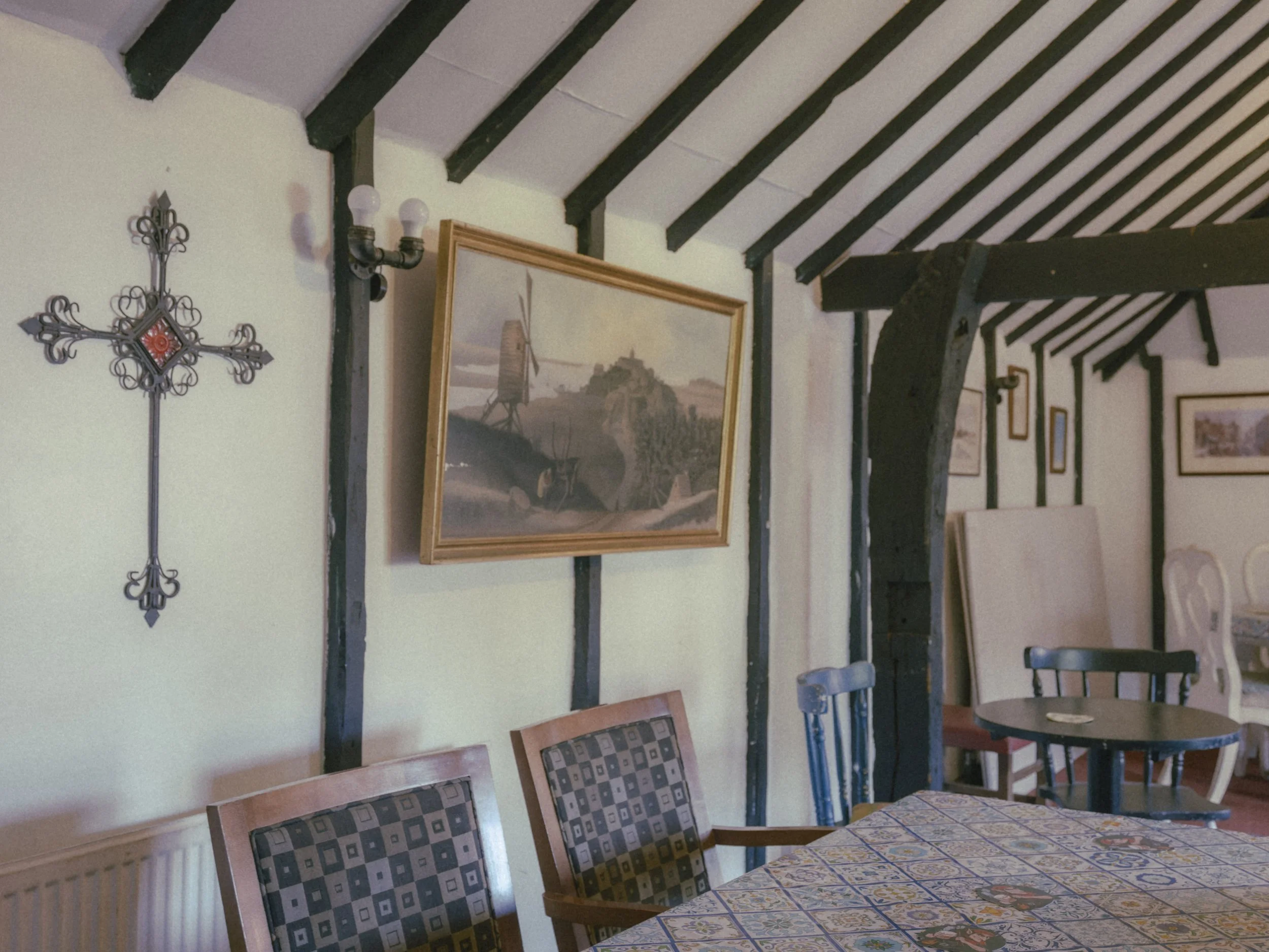 A dining room with black exposed wooden beams on the ceiling, framed artwork depicting a windmill and rural landscape, a metal wall sconce with two light bulbs, a row of chairs with patterned upholstery, a table with a colorful tiled tablecloth, and 