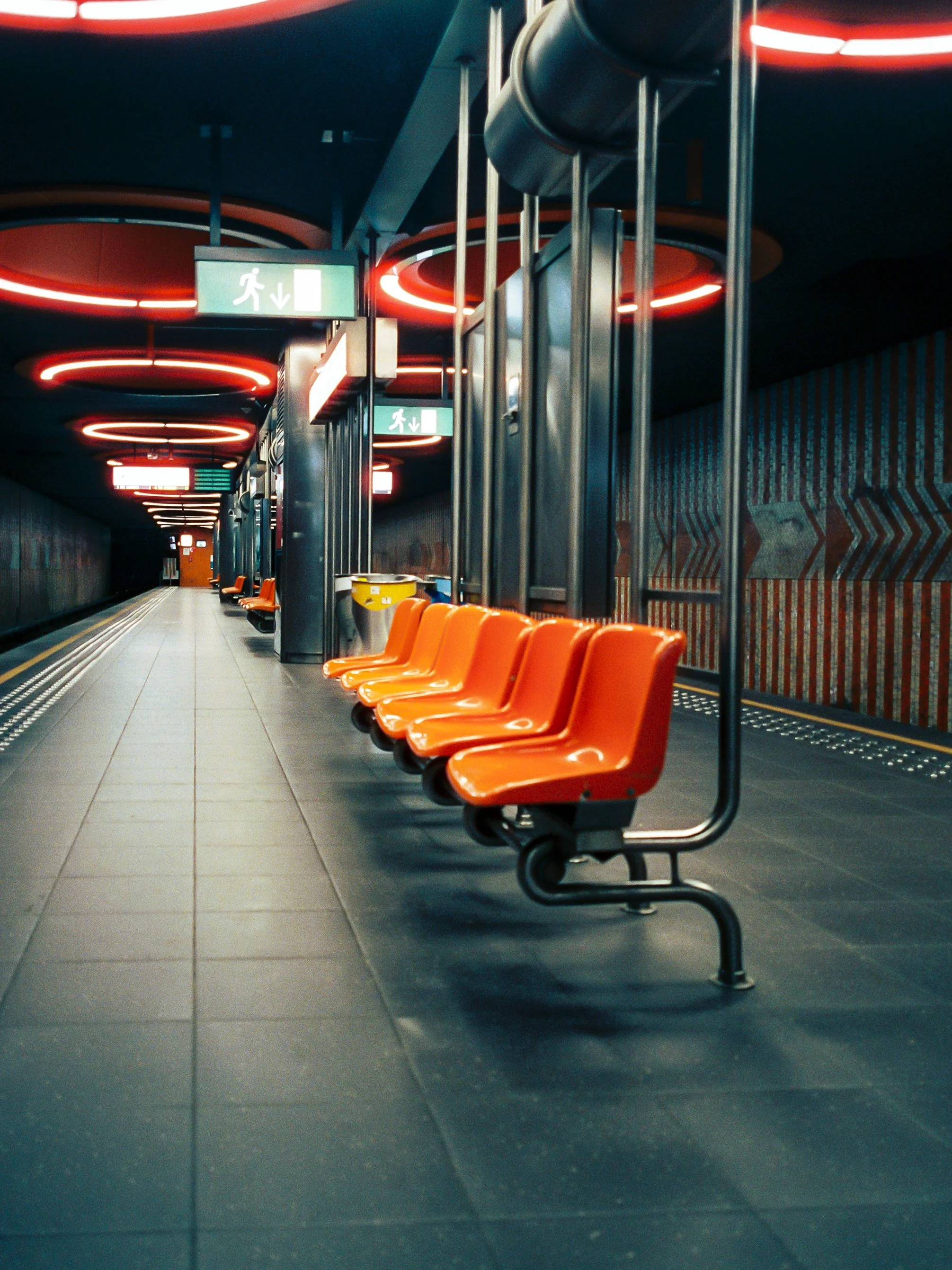 Empty orange seats in a subway station with a digital display and exit signs overhead, featuring modern dark interior design with red circular ceiling lights.