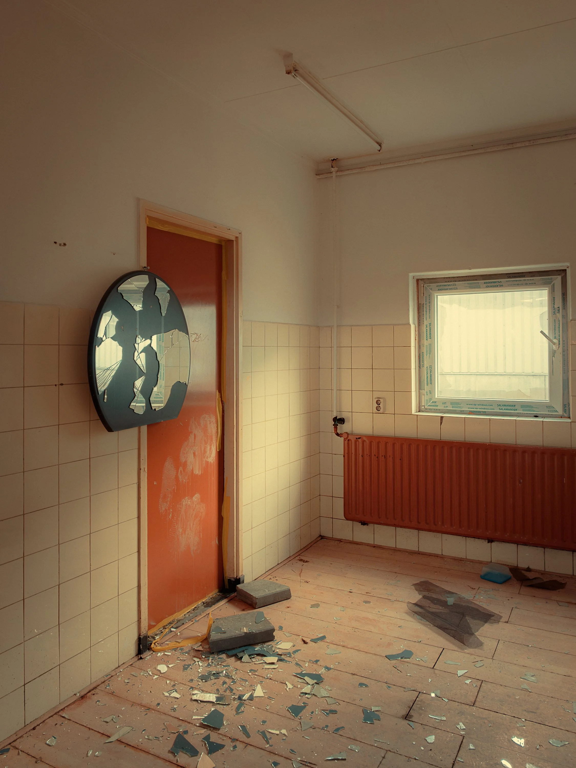 A room with broken mirror pieces scattered on the wooden floor, a damaged mirror hanging on the wall, a window with a partly open blind, and an orange door with some paint damage. The ceiling has exposed wiring and a light fixture. A radiator is moun