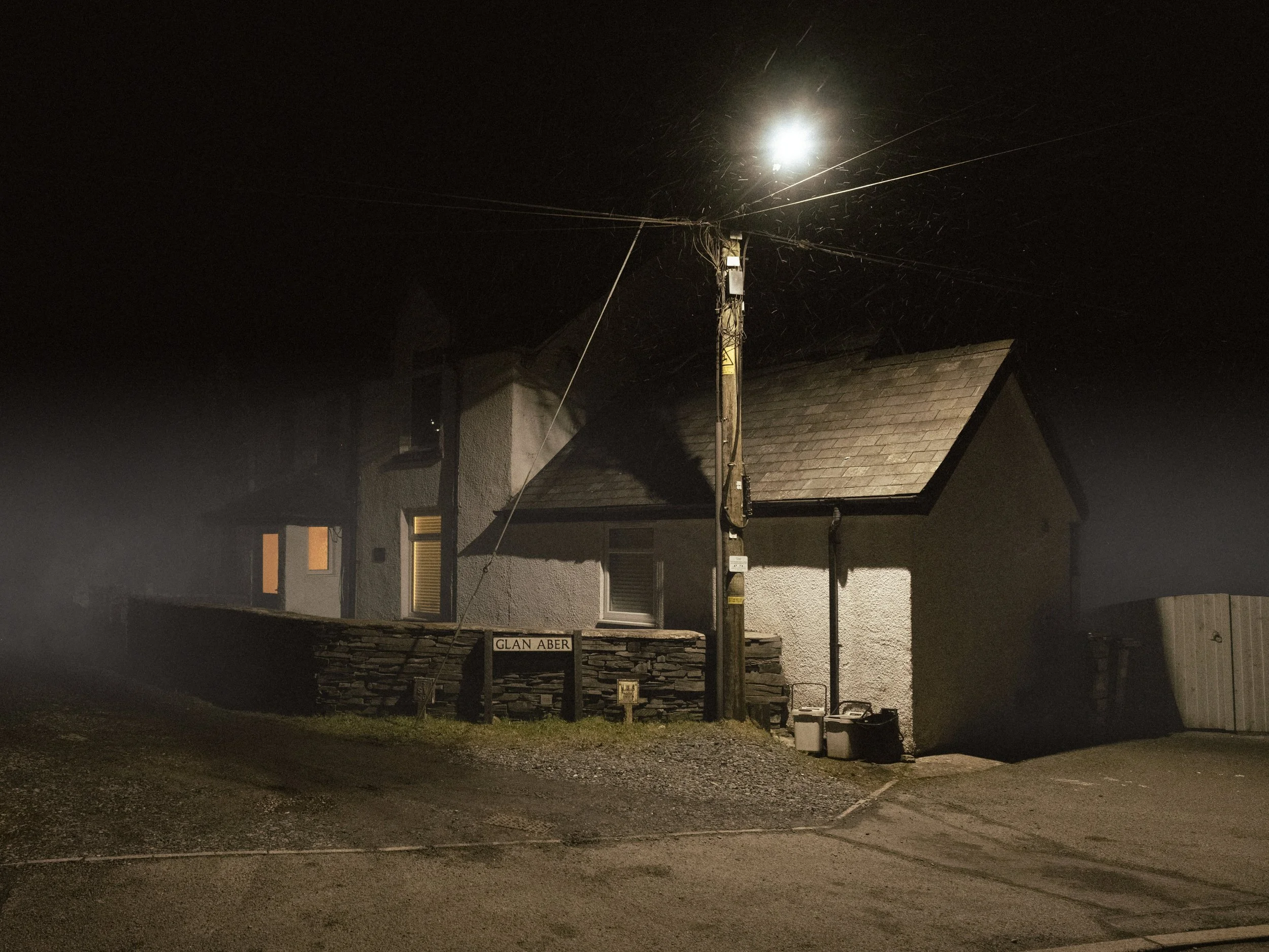 Nighttime scene of a house with a streetlamp illuminating the area. There is a street sign labeled "Glan Aber" in front of the house, and the roof of the house is visible. The street appears empty and quiet.