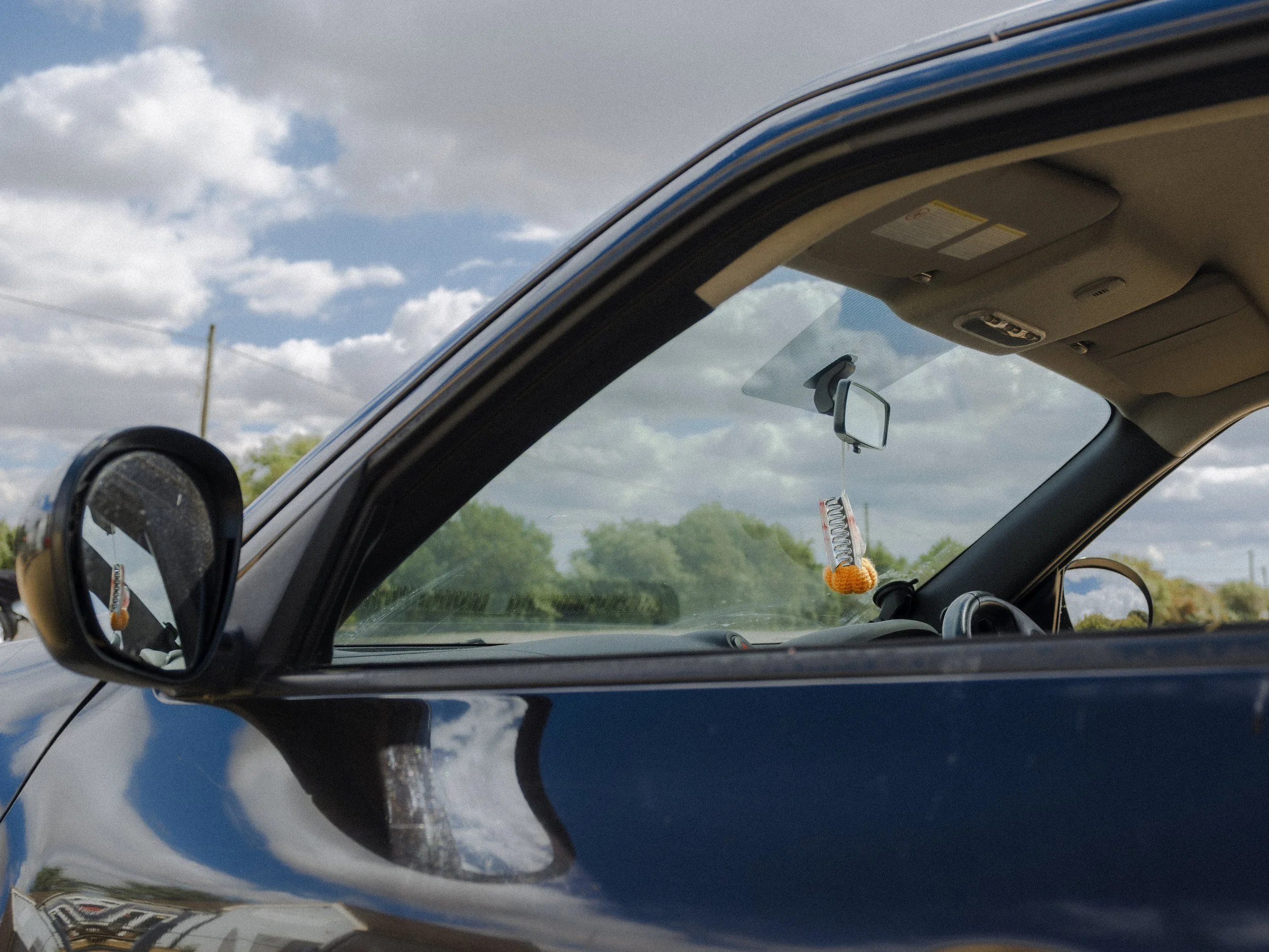 View of the interior dashboard and driver-side window of a vehicle with a religious or decorative item hanging from the rearview mirror, showing a cloudy sky outside.
