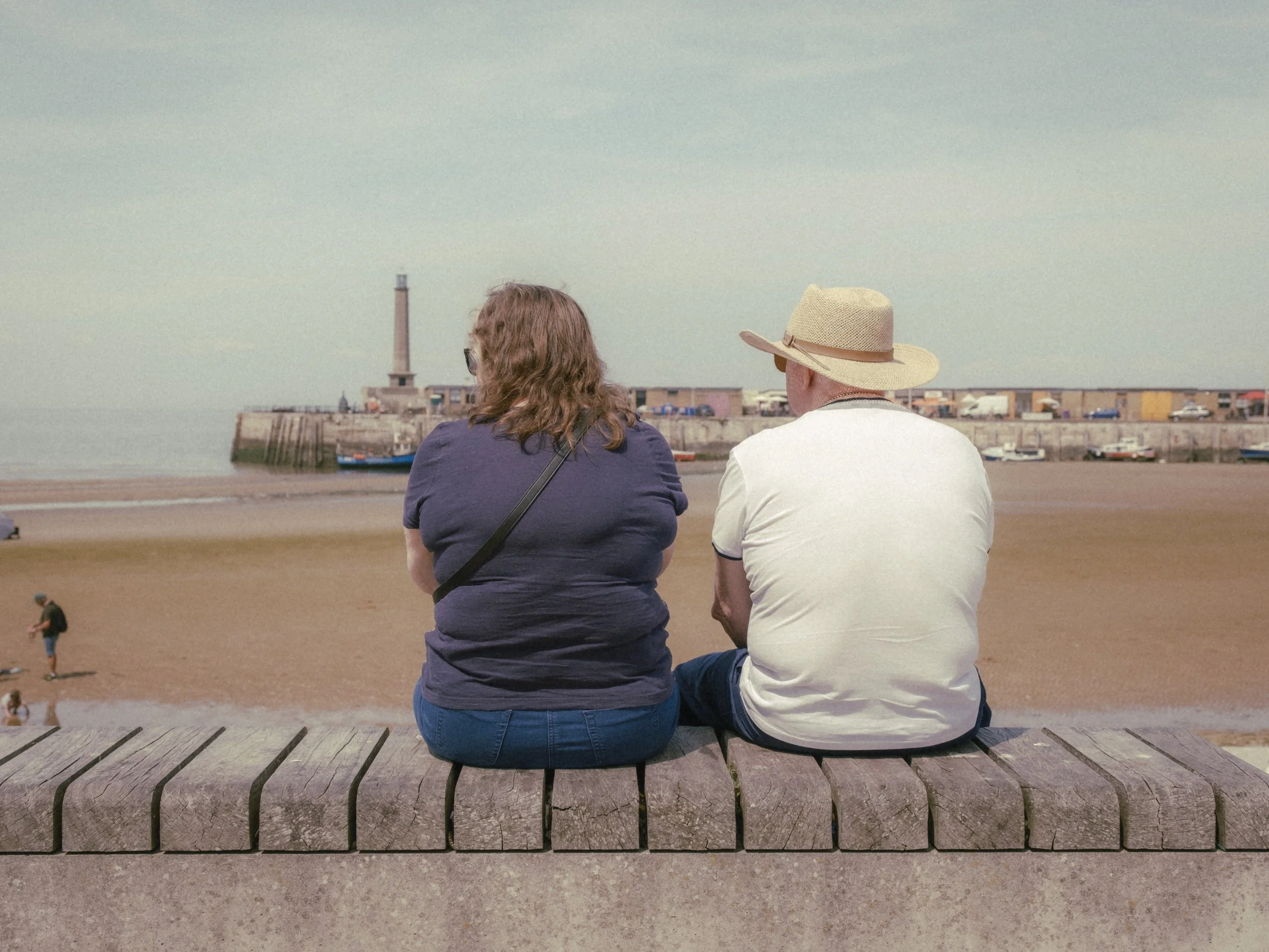 Two people sitting on a bench at the beach, facing the ocean, with a lighthouse and a pier in the background.