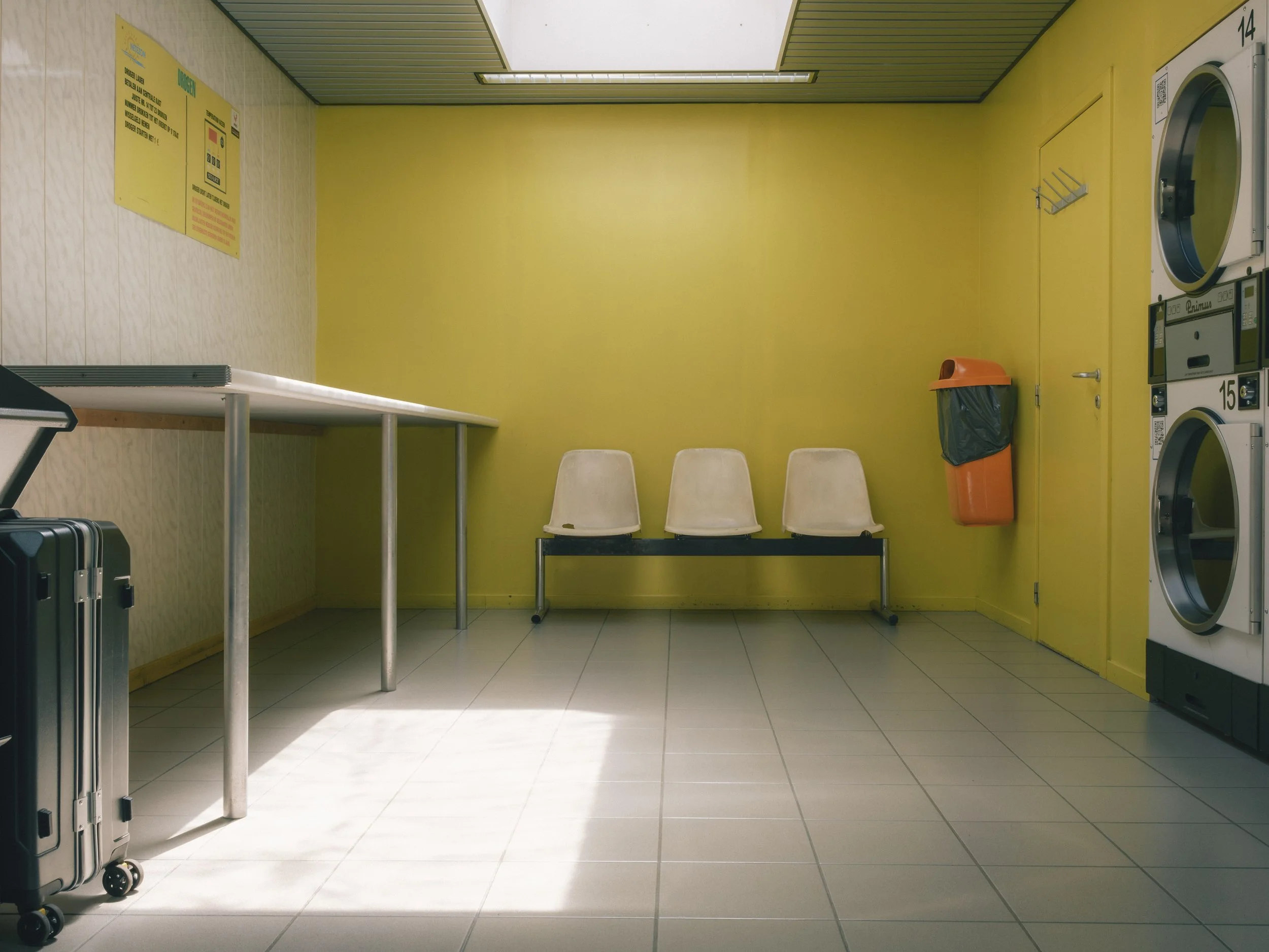 Laundry room with yellow walls, white chairs, a gray table, a rolling suitcase, a washing machine, a dryer, and a trash bin.