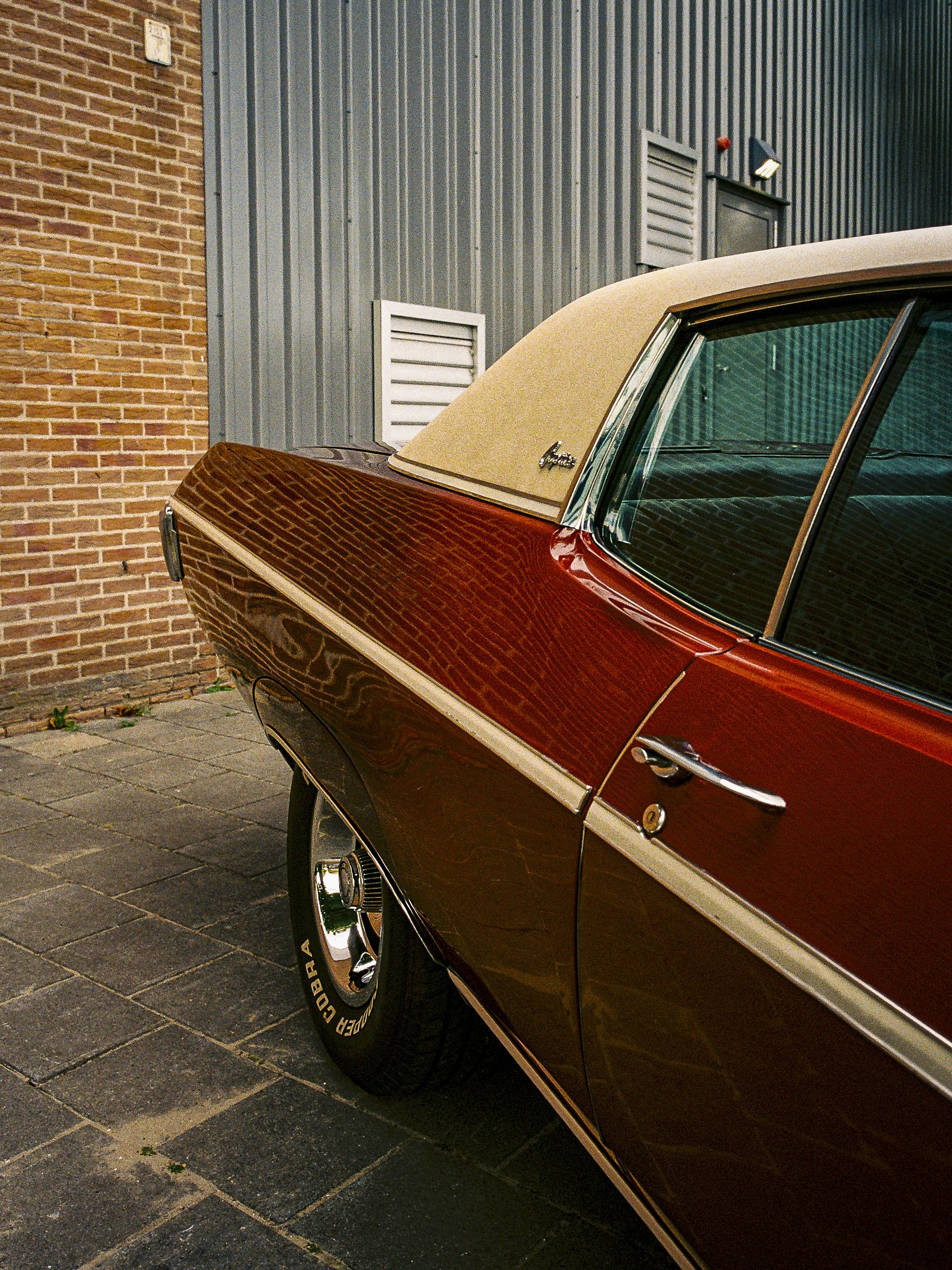 Close-up of the rear side of a vintage red and beige car parked on a brick and concrete sidewalk, with a gray metal building in the background.