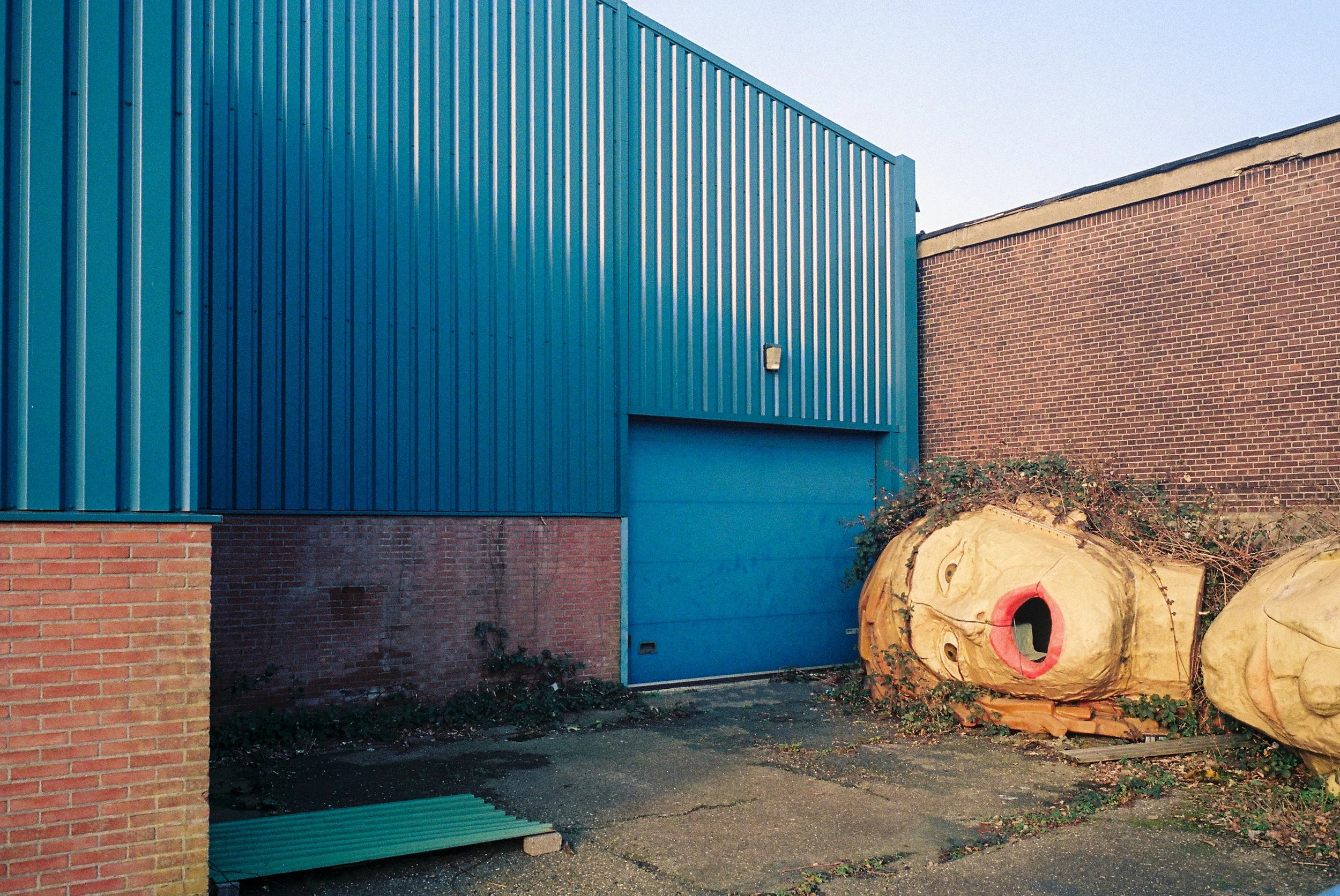 Blue metal building with a brick wall section on the left, a light fixture on the blue wall, and several large tree logs with a face painted on one log, placed on the ground in front of the building.