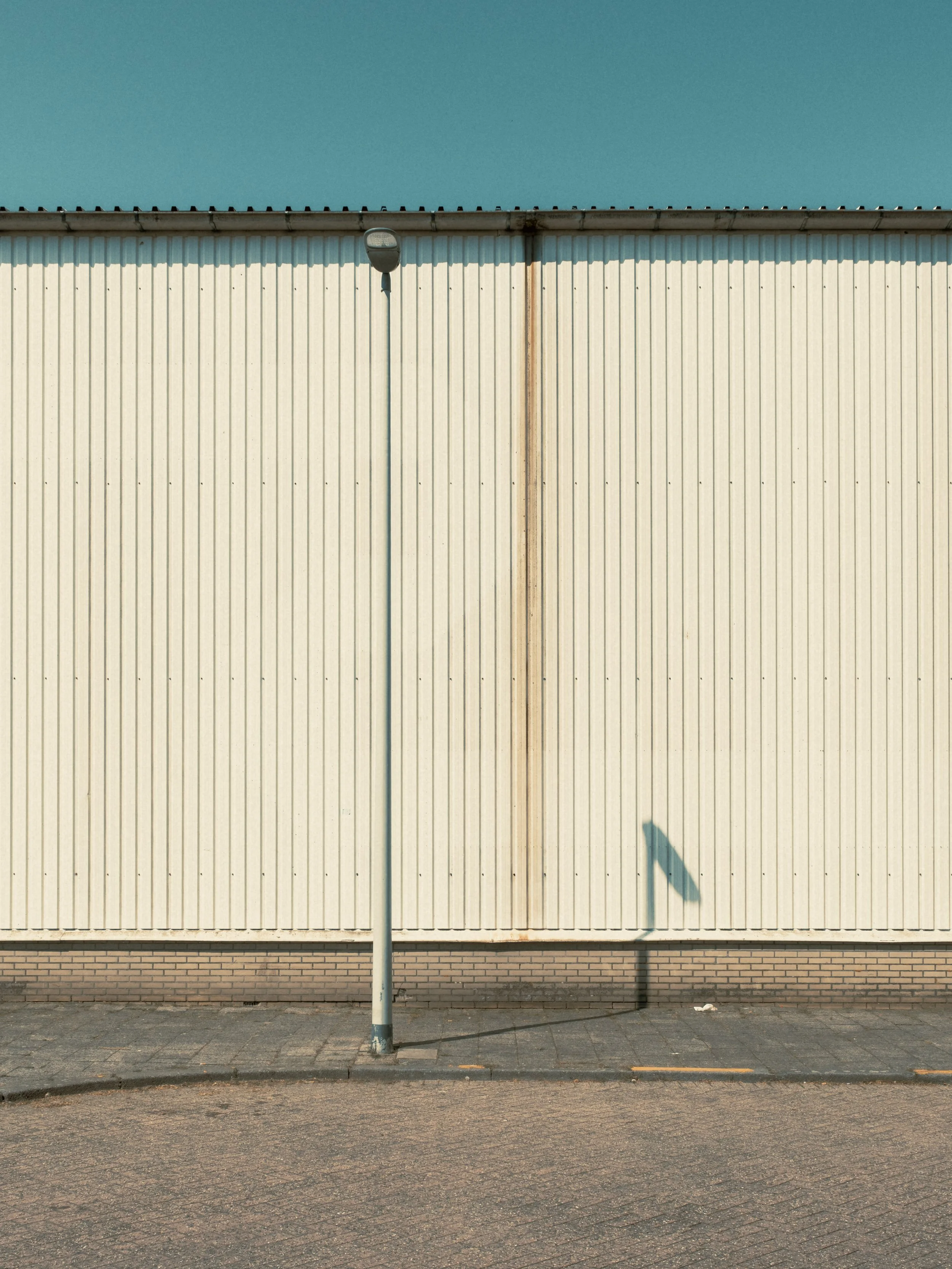 A tall street light in front of a beige metal building with vertical ridges. The sky is clear and blue, and there are shadows cast on the ground by the streetlight and a small object near it.