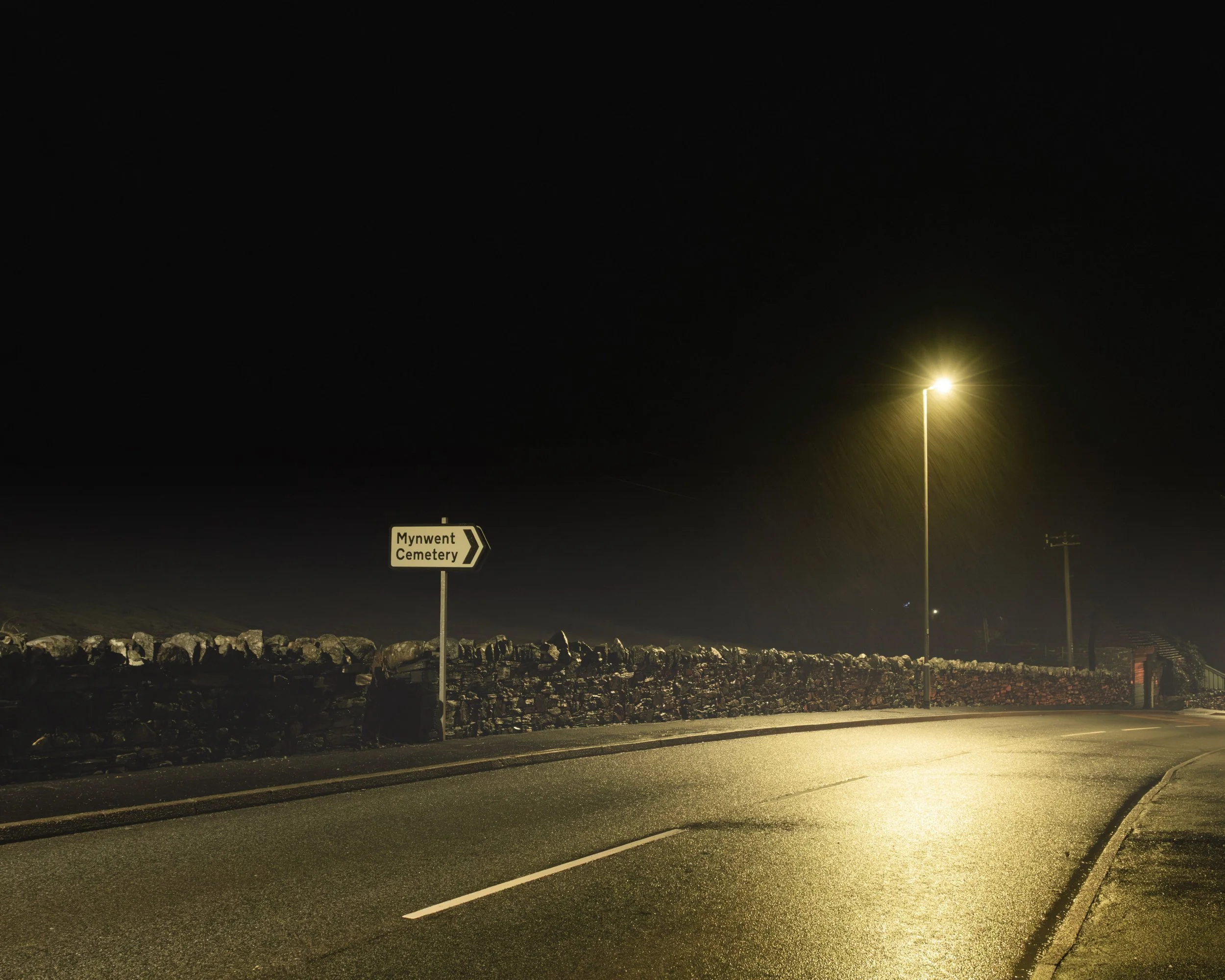Nighttime scene with a streetlamp illuminating a road with a sign pointing to Mynwent Cemetery. The sky is dark and clear, with rocks and pavement visible in the foreground.