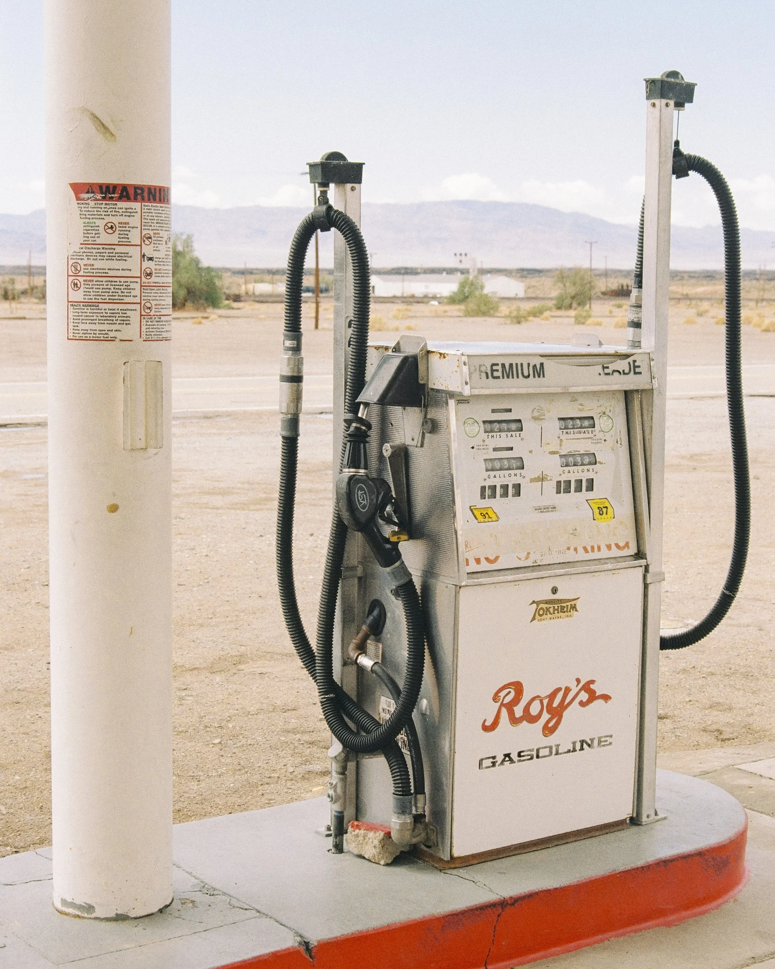 Gas pump with hoses and meters, next to a white pole, in a desert area with mountains in the background.