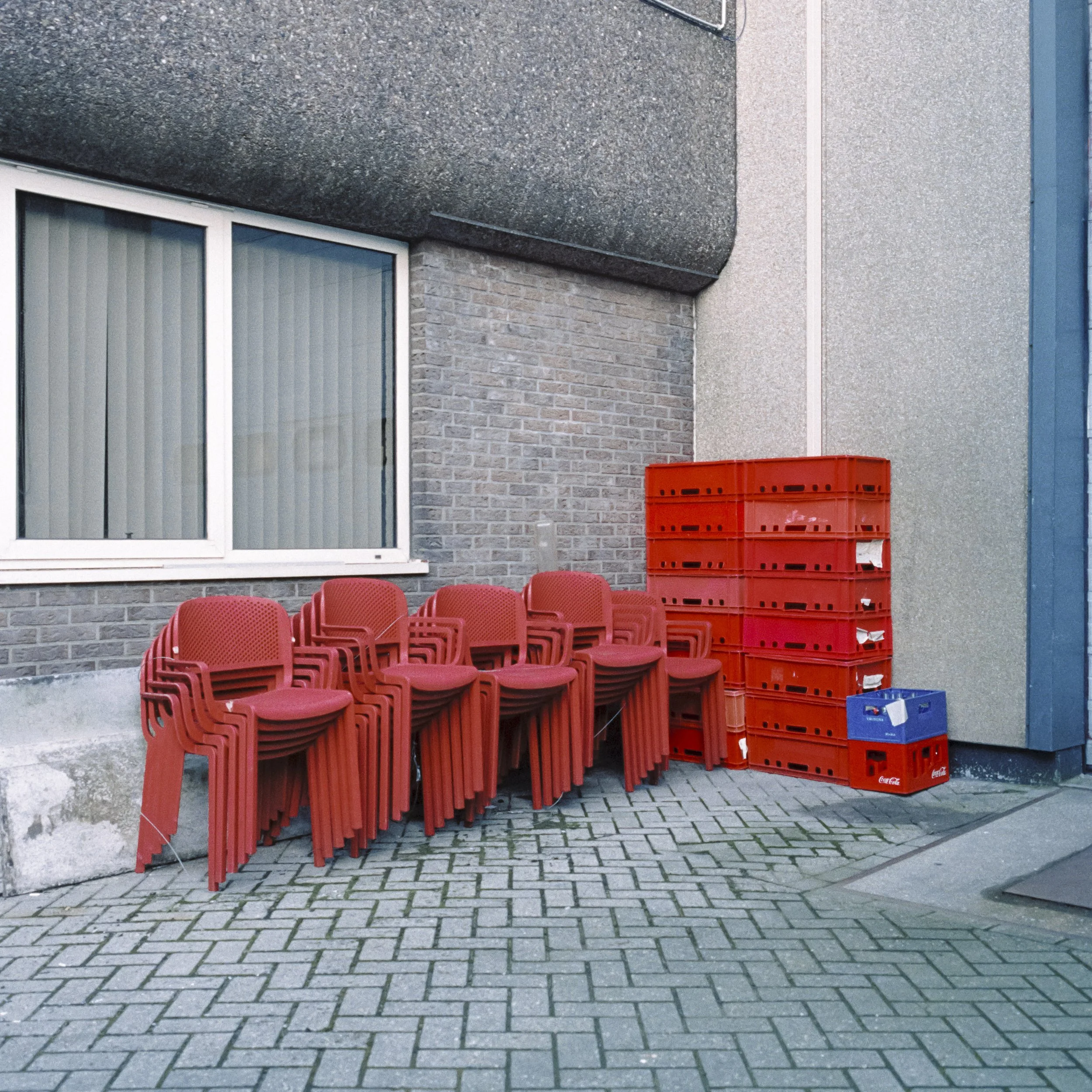 Red plastic crates stacked outside a building with gray brick wall, glass window, and concrete pavement.