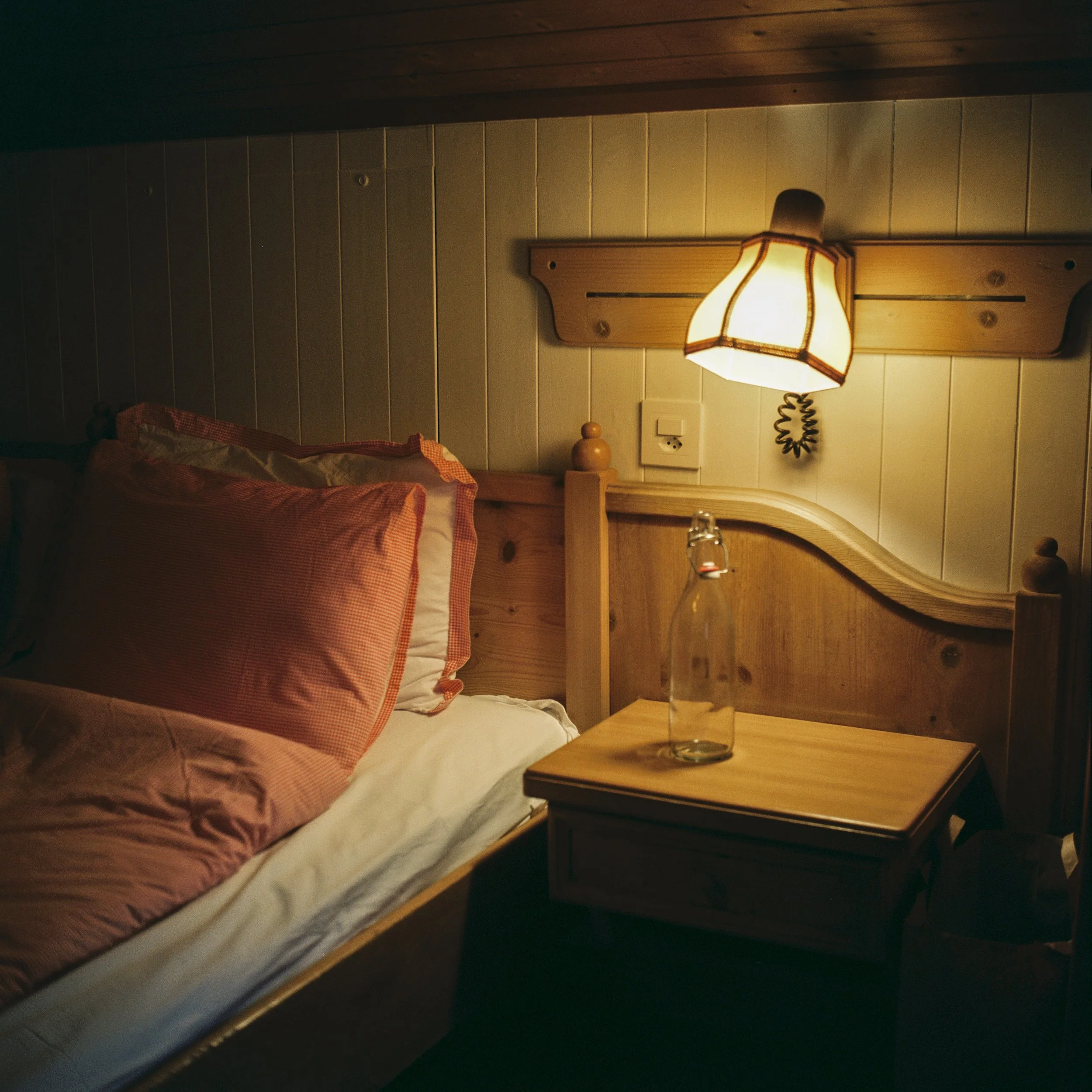 A cozy bedroom corner with a wooden bed, a bedside table with a glass of water, and a wall-mounted lamp casting warm light.